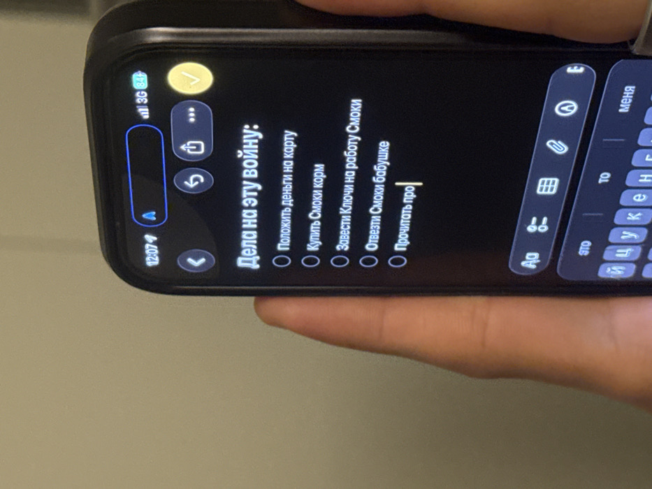 The scene is located in Tel-Aviv, Israel. A human hand, exhibiting a light to medium skin tone, is holding a dark-colored smartphone, likely an iPhone, encased in a dark protective cover. The phone's screen displays a task list application in a dark mode interface, viewed from a slight angle. The top left of the screen shows the time as 12:07, and the top right indicates 3G network connectivity and an 84% battery charge.
The title of the task list, prominently displayed in Russian, reads "Дела на эту войну:" (Tasks for this war:). Below the title, five tasks are listed, each preceded by an unchecked circular checkbox:
1. "Положить деньги на карту" (Deposit money onto the card)
2. "Купить Смоки корм" (Buy Smokey's pet food)
3. "Завести Ключи на работу Смоки" (Bring keys to Smokey's work)
4. "Отвезти Смоки бабушке" (Take Smokey to grandma)
5. "Прочитать про|" (Read about|), with a blinking cursor indicating active text input.
A Cyrillic keyboard is partially visible at the bottom portion of the screen. The background is a plain, out-of-focus, light-colored wall.