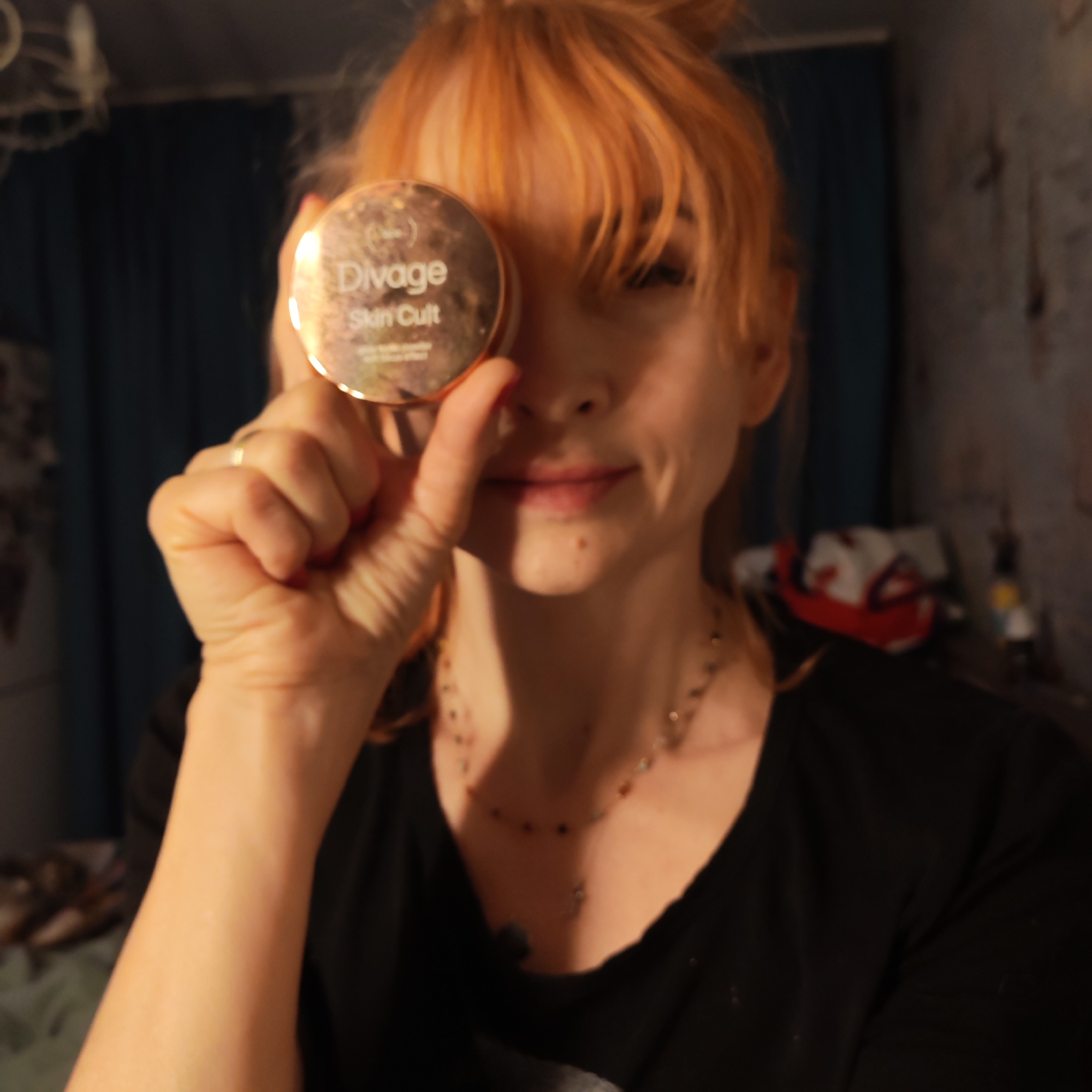 A woman with reddish-orange hair is holding a round, gold-colored compact labeled "Divage Skin Cult" in front of her right eye. She is wearing a black shirt and a multi-colored necklace. The background is slightly blurred but appears to be an interior room with patterned curtains and various objects on a table or surface behind her. The location may be in a city of Chaikovsky, Russia.