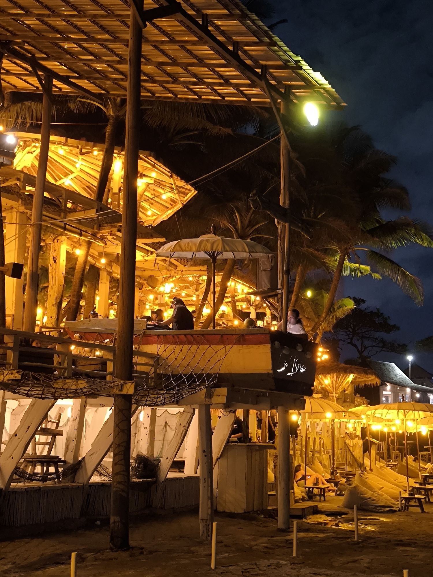 The image depicts a dimly lit outdoor dining area at night, potentially located in Canggu, Indonesia, based on the architectural style and atmosphere. The scene is dominated by a multi-level wooden structure, likely a restaurant or bar, decorated with numerous warm-toned string lights. People are seated at tables on the upper level of the structure, partially obscured by an umbrella.
Below, the ground level shows additional seating arrangements with bean bags and low tables. The overall setting suggests a tropical or beachside environment with visible palm trees. The structure has a weathered appearance, indicative of coastal architecture. A sign with stylized writing is mounted on the side of the upper level. The lighting creates a warm, inviting ambiance.