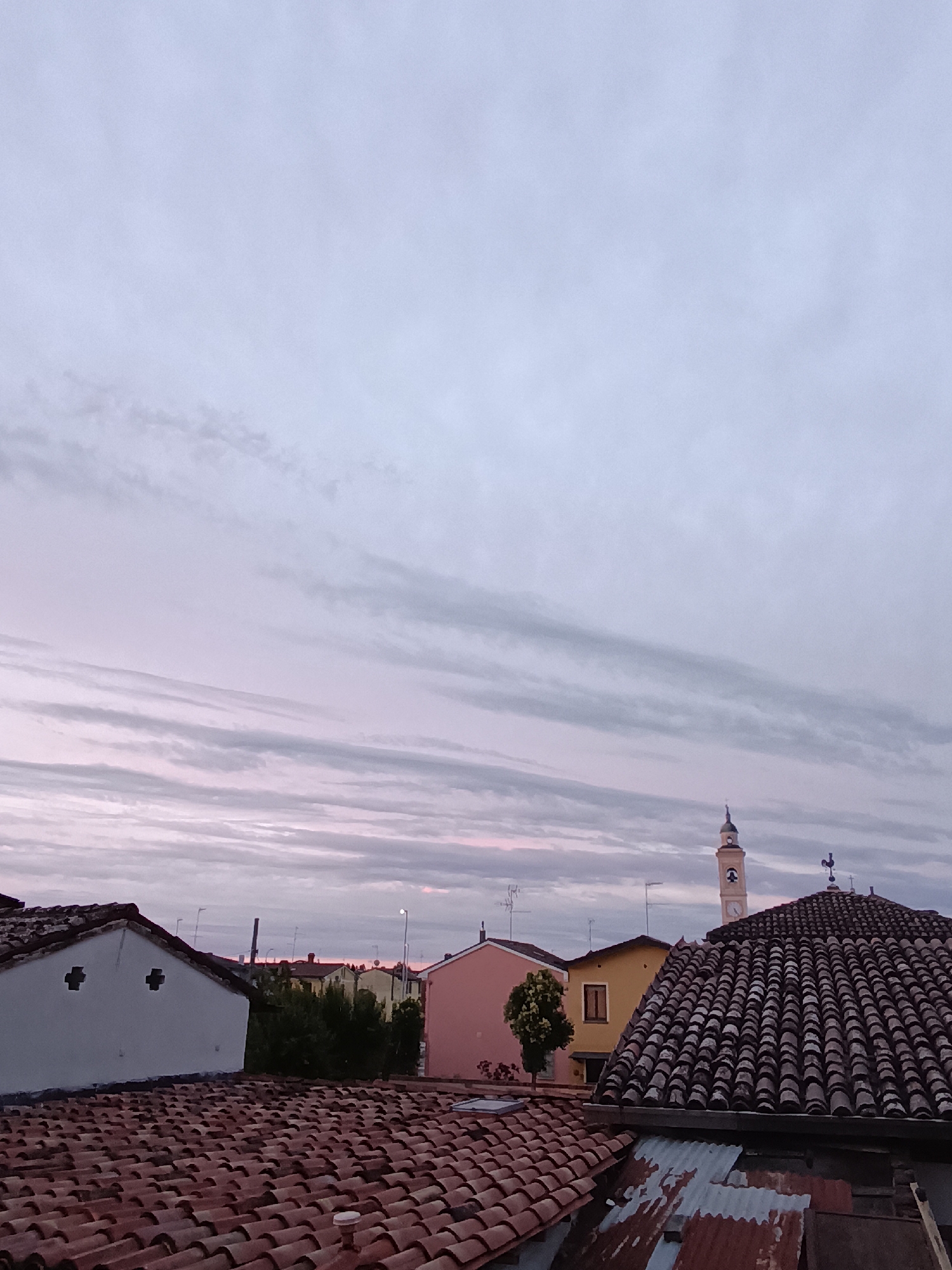 This image depicts a view of rooftops and a distant church tower under a cloudy, twilight sky in Viadana, Italy. The foreground is dominated by a large expanse of terracotta roof tiles, some appearing aged and weathered. To the left, a white building with two distinct cross-shaped window openings is visible, also featuring a tiled roof. Beyond these structures, several other buildings are present, including a pink house, a yellow house, and behind them, a church with a distinctive bell tower and a green-domed spire. The sky is filled with long, streaky clouds that are tinged with pink and purple hues, indicating either sunrise or sunset. There are no people visible in the image. The overall impression is of a quiet, residential area in an Italian town at dusk.