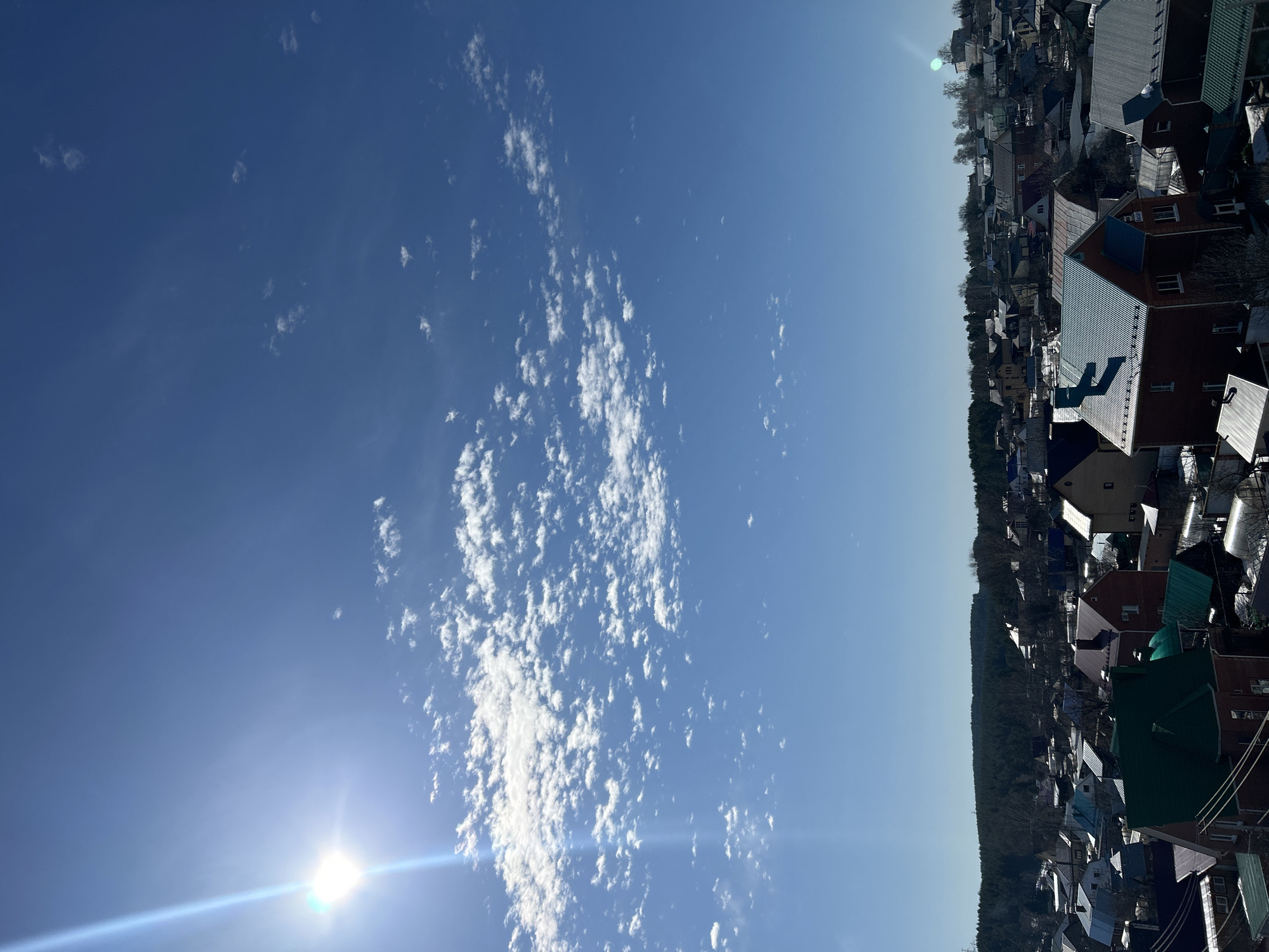 The image depicts a view over the city of Chaikovsky, Russia, under a bright blue sky with scattered altocumulus clouds. The sun shines brightly, creating a lens flare at the bottom left of the frame. The cityscape appears to consist of densely packed residential buildings, likely houses and small apartment buildings with varied roof colors and styles. A line of trees can be seen in the mid-ground, likely outlining a park or natural area within the city. The photograph is taken from an elevated vantage point.