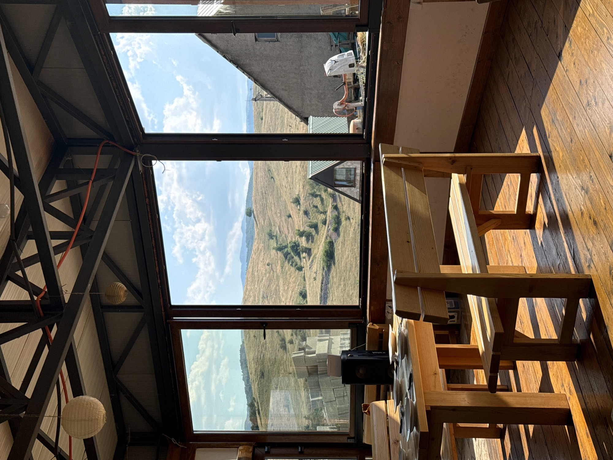 An interior view of a rustic dining area with large windows overlooks a sun-drenched, rolling landscape. The room features wooden tables and benches, with sunlight casting sharp shadows across the polished wooden floorboards. In the foreground, a wooden table is set with plates and cutlery. Beyond the table, a wooden bench with slatted seating and backrest is visible. The expansive windows offer a view of a dry, grassy hillside dotted with trees and a few buildings. In the middle distance, a small, dark-colored speaker sits on the table. The ceiling is a mix of exposed metal beams and wooden panels, with two paper lanterns hanging from above. The scene appears to be located in Rudinice, Montenegro.