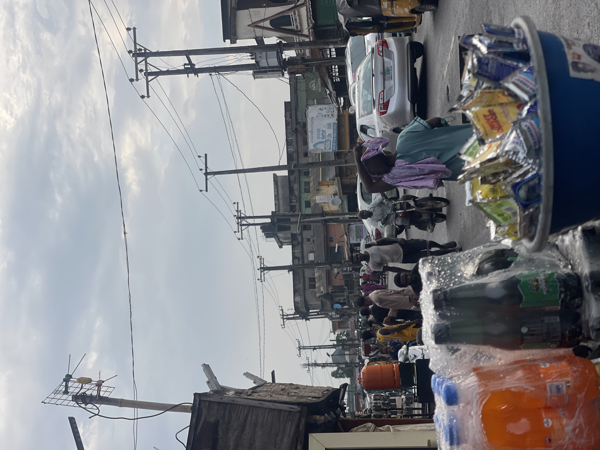 The image depicts a street scene in Akowonjo, Nigeria, characterized by its bustling atmosphere and infrastructure typical of a commercial area. Overhead, a dense network of electrical wires and poles crisscrosses the sky, indicating a well-established power grid. The street itself is populated by a variety of people, some walking, others on motorcycles or in cars, suggesting a lively commercial district. A prominent feature is a collection of bottled drinks wrapped in plastic, placed in the foreground, likely for sale, along with a blue bucket filled with various packaged goods. Buildings line the street, displaying a mix of architectural styles and ages, with some appearing to be shops or businesses. Vehicles, including cars, motorcycles, and a three-wheeled auto-rickshaw, are present, contributing to the sense of an active urban environment. The overall impression is one of a vibrant marketplace with daily life unfolding under a cloudy sky.