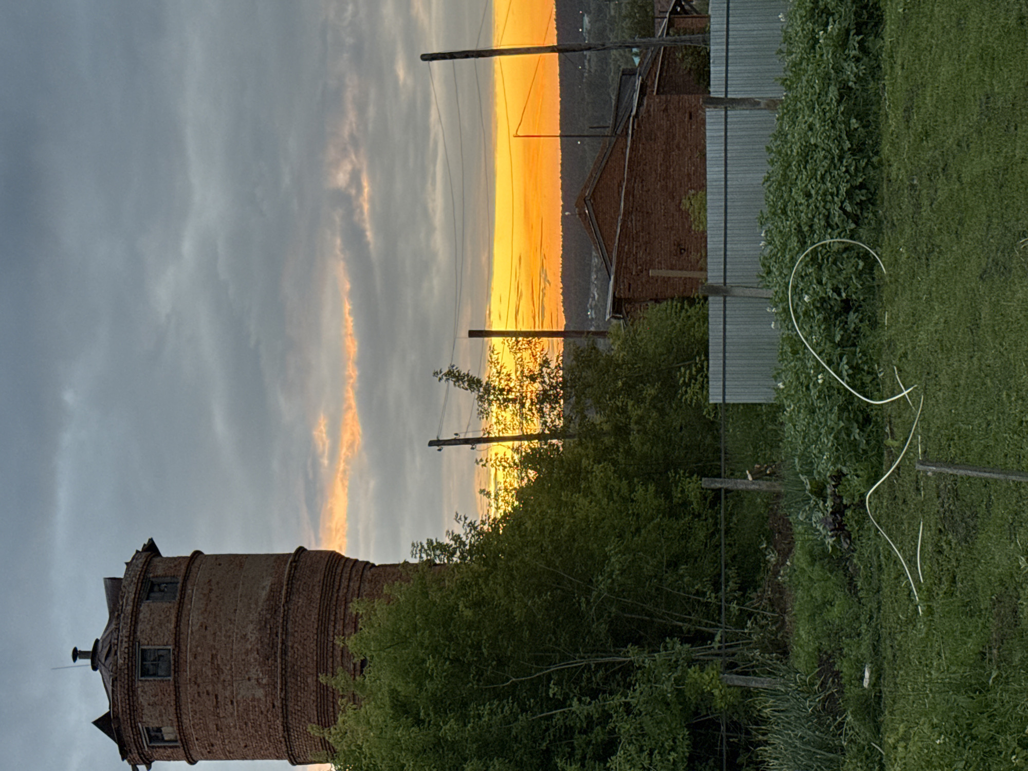 This image captures an exterior scene at dusk, likely in or near the city of Malmyzh, Russia. Dominating the left side of the frame is a tall, weathered brick water tower, its cylindrical structure showing signs of disrepair with broken windows and a damaged roof. A dense canopy of green trees partially obscures the tower, reaching from the foreground towards its base.
In the background, a vibrant sunset paints the sky with intense shades of orange and yellow, with softer hues of pink and gray clouds interspersed. Silhouetted against this dramatic sky are power lines supported by wooden poles, suggesting the presence of infrastructure. Below the sunset, a line of dark trees forms the horizon, with glimpses of buildings in the distance.
Closer to the viewer, a red brick building with a dark roof is visible. In the foreground, a grassy area transitions into a garden or cultivated plot with rows of green plants, possibly potatoes, exhibiting small white flowers. A light-colored hose is coiled on the ground in the garden. A simple fence, constructed from wooden posts and corrugated metal sheeting, runs across the mid-ground, separating the garden from the area with the brick building. The overall atmosphere is tranquil and rural, with the natural beauty of the sunset contrasting with the rustic, somewhat neglected man-made structures. There are no people visible in the scene.
