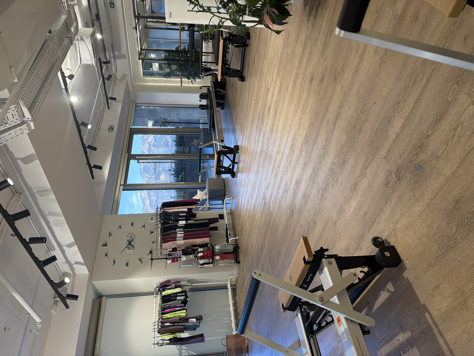 The image depicts the interior of a modern Pilates studio, likely located in Boreti, Montenegro, given the scenic mountain and sea views visible through the large windows. The studio features polished wooden floors and an abundance of natural light. In the foreground, a Pilates reformer machine is prominently displayed, with its metal frame and wooden accents.
Further into the studio, more Pilates reformers are arranged, suggesting a space designed for group or individual training sessions. The walls are adorned with minimalist artwork, including a clock with silhouette figures engaged in Pilates poses. Several clothing racks filled with activewear are visible on the left side of the image, indicating that the studio may also sell apparel.
The large windows offer a breathtaking panoramic view of a mountainous coastline, hinting at the studio's picturesque location in Boreti. The overall ambiance is one of clean lines, functionality, and a connection to the natural surroundings, creating a serene and inviting environment for wellness activities. There are no people visible in the image.