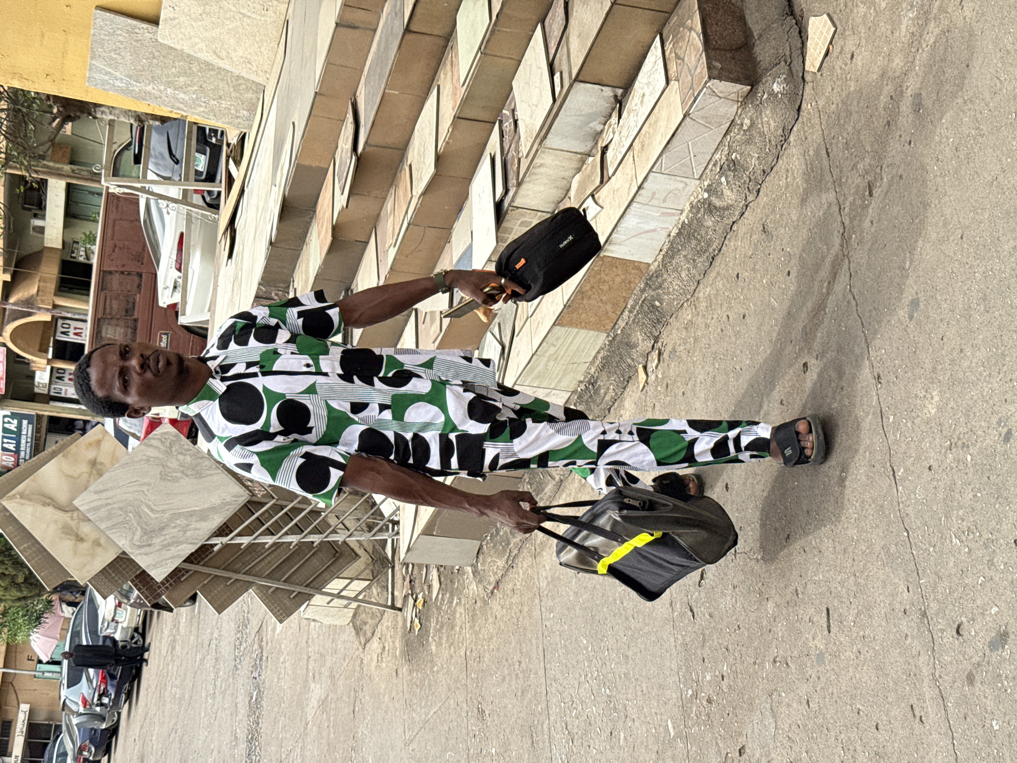 A man stands on a sidewalk near a building entrance with stairs tiled in a variety of patterns. He wears a patterned, green, black, and white shirt and pants, and slides on his feet. He holds a black bag with yellow trim in one hand and a smaller black bag in the other. Tile samples in a metal rack are visible to the left of the man. Text on the building sign reads "TANI BUSINESS RACHINE JANA BIZ MACHINE AO A1 A2", "OV", "LV", "food", and "HurleyX". Cars are parked along the street in the background, and the image appears to be taken in Abuja, Nigeria.