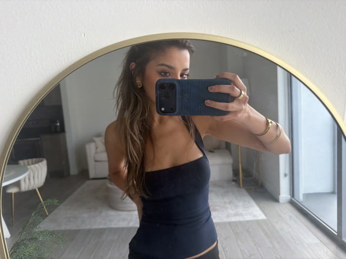 An adult female is captured in a mirror selfie within what appears to be a modern apartment or condominium. She stands facing a large, circular mirror with a thin gold-colored frame, which is mounted on a light-colored wall. The female has long, wavy, light brown hair, parted to one side, and wears a dark-colored, one-shoulder tank top. She holds a dark-colored smartphone, encased in a textured dark blue case, with her left hand, obscuring part of her face as she photographs her reflection. Her left hand displays a gold-colored ring on her ring finger, and her left forearm is adorned with multiple thin, gold-colored bangles. A gold-colored earring is visible in her right ear.
The reflection in the mirror reveals an open-plan living space with contemporary decor. To the left, a round white table is paired with a ribbed, light-colored chair featuring gold-colored legs. A small green plant is partially visible near the base of the mirror. In the central background, a light-colored armchair and a round ottoman are present, situated on a light-colored rug covering a section of the light wood-toned floor. Further back, a dark kitchen counter with appliances is partially visible. To the right, a large glass sliding door or window provides a glimpse of an outdoor balcony area, suggesting an urban residential setting. The overall environment indicates a well-lit, minimalist interior.
The architecture and interior design, featuring modern finishes and an outward-facing balcony, are consistent with high-rise residential buildings commonly found in urban areas, including within the city of Miami, United States of America. No specific landmarks identifying Miami are visible in the image.