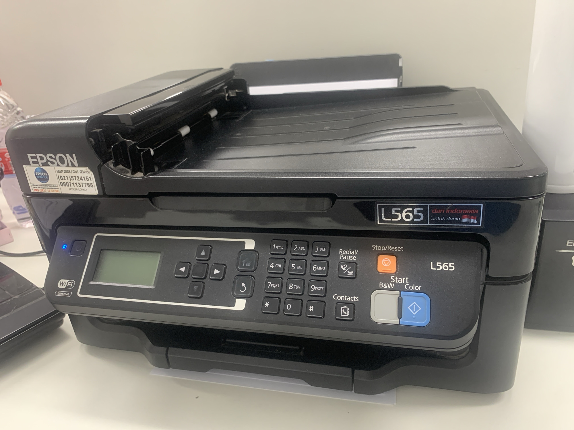 This is a medium shot, eye-level, indoor photograph of an Epson L565 all-in-one printer. The printer is black and is positioned on a white desk surface. The top lid of the printer is open, revealing the scanner mechanism. To the left of the scanner is an Epson service sticker with contact information and a bottle of what appears to be liquid. To the right of the scanner, a white cylindrical object is partially visible. The printer's control panel is visible and features a digital display, a numeric keypad, navigation buttons, and function buttons including "Stop/Reset", "Start B&W", and "Color". A blue light is illuminated next to the power button, and a Wi-Fi/Ethernet icon is present. The printer is located on a desk in an office environment. There is no indication of the specific location within Palembang, Indonesia.