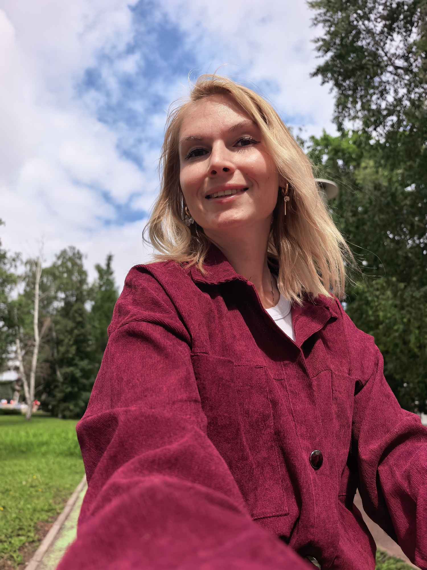 A blonde-haired woman, approximately 20-30 years old, with fair skin and brown eyes, smiles directly at the camera, capturing a close-up selfie in Sterlitamak, Russia. She wears a dark maroon, corduroy-textured jacket over a white t-shirt, featuring a single visible dark button on the front and gold-colored dangling earrings. The outdoor setting under a clear blue sky with white clouds reveals a park-like environment. The background comprises numerous trees, including light-barked trees and dense green foliage, with a grassy area and a paved path or curb with a green edge visible in the lower left. Distant urban elements are faintly discernible through the trees on the far left.
