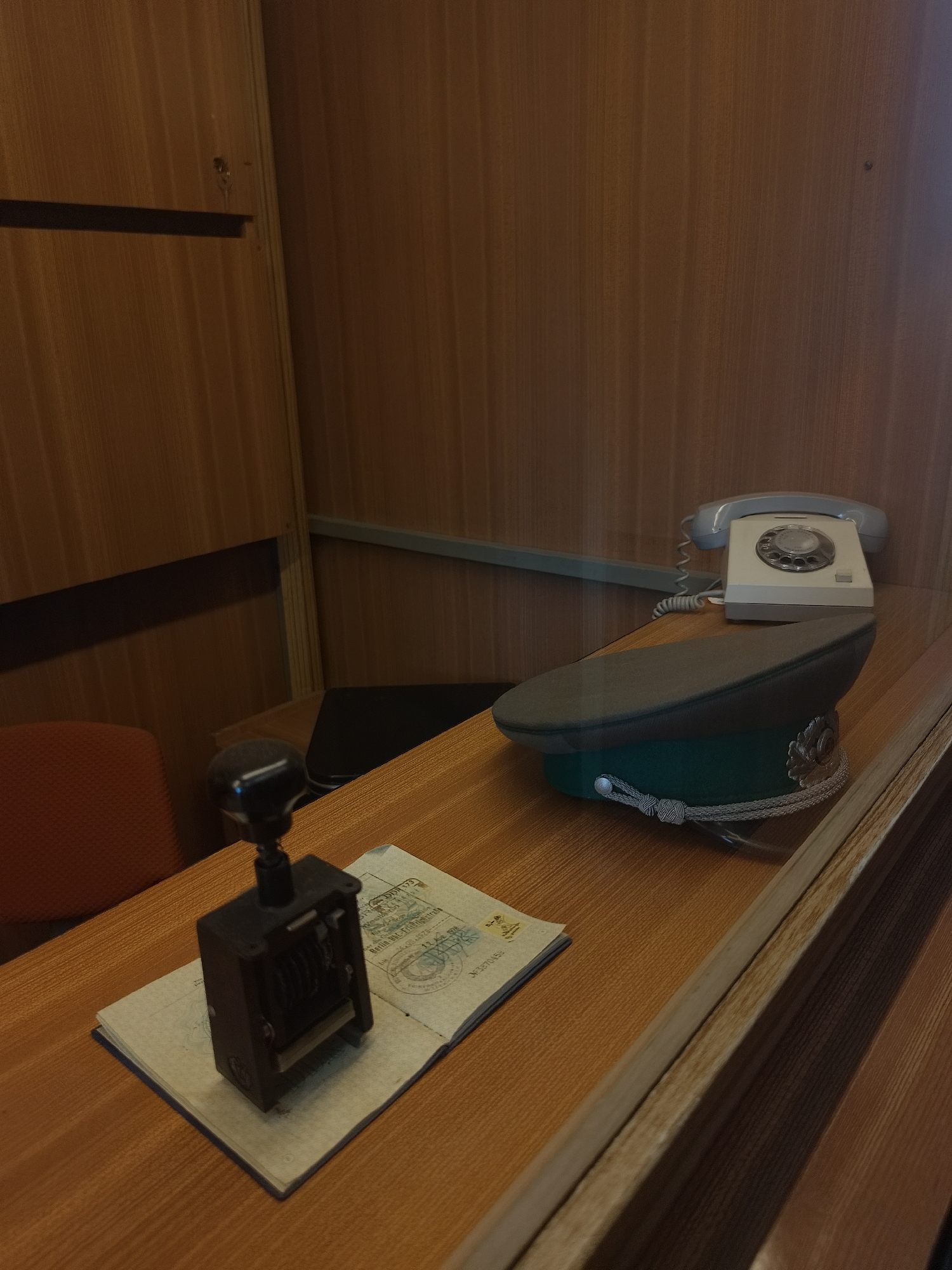 The image depicts a scene likely set in a museum or a historical exhibit, presenting a mock-up of a border checkpoint or an official's office. The focus is on the objects on the desk, which include a black stamp on top of an open passport, an officer's cap, and a vintage rotary telephone. The background consists of wooden paneling, suggesting an enclosed space. The lack of people and the static nature of the objects indicates a staged setting. The elements suggest a place where official documents are processed, possibly at a border crossing or immigration office, but without a specific location, its location in the city of Unknown, Unknown cannot be verified.