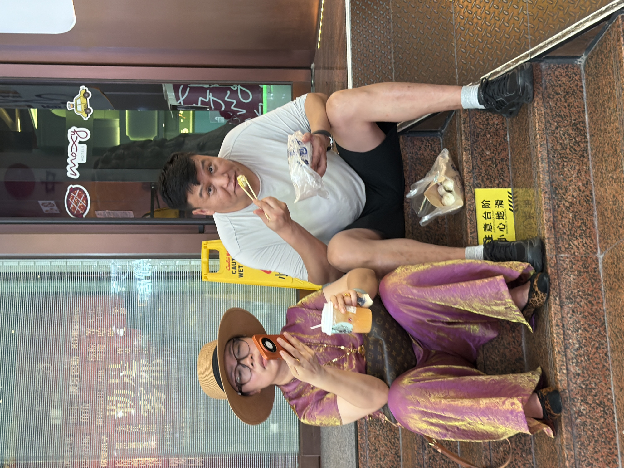 The image depicts two individuals sitting on a set of granite steps in front of a building. On the left, a person wearing a wide-brimmed straw hat, glasses, a purple and gold iridescent outfit, and leopard print flats holds an orange cell phone to their ear and has a cup of amber-colored liquid with a straw in their other hand. A brown designer handbag with a monogram pattern is slung over their shoulder. To the right, a man wearing a light grey t-shirt and black shorts, with black athletic shoes and grey socks, is eating from a plastic bag of food with chopsticks. A yellow caution sign with Chinese characters and "CAUTION WET" in English is visible behind the woman. The building behind them has a glass facade with horizontal blinds and displays some stickers on the door, including one that reads "moxy HOTELS". The overall setting appears to be an urban environment in Yuzhong District, China, likely an entrance to a commercial establishment.