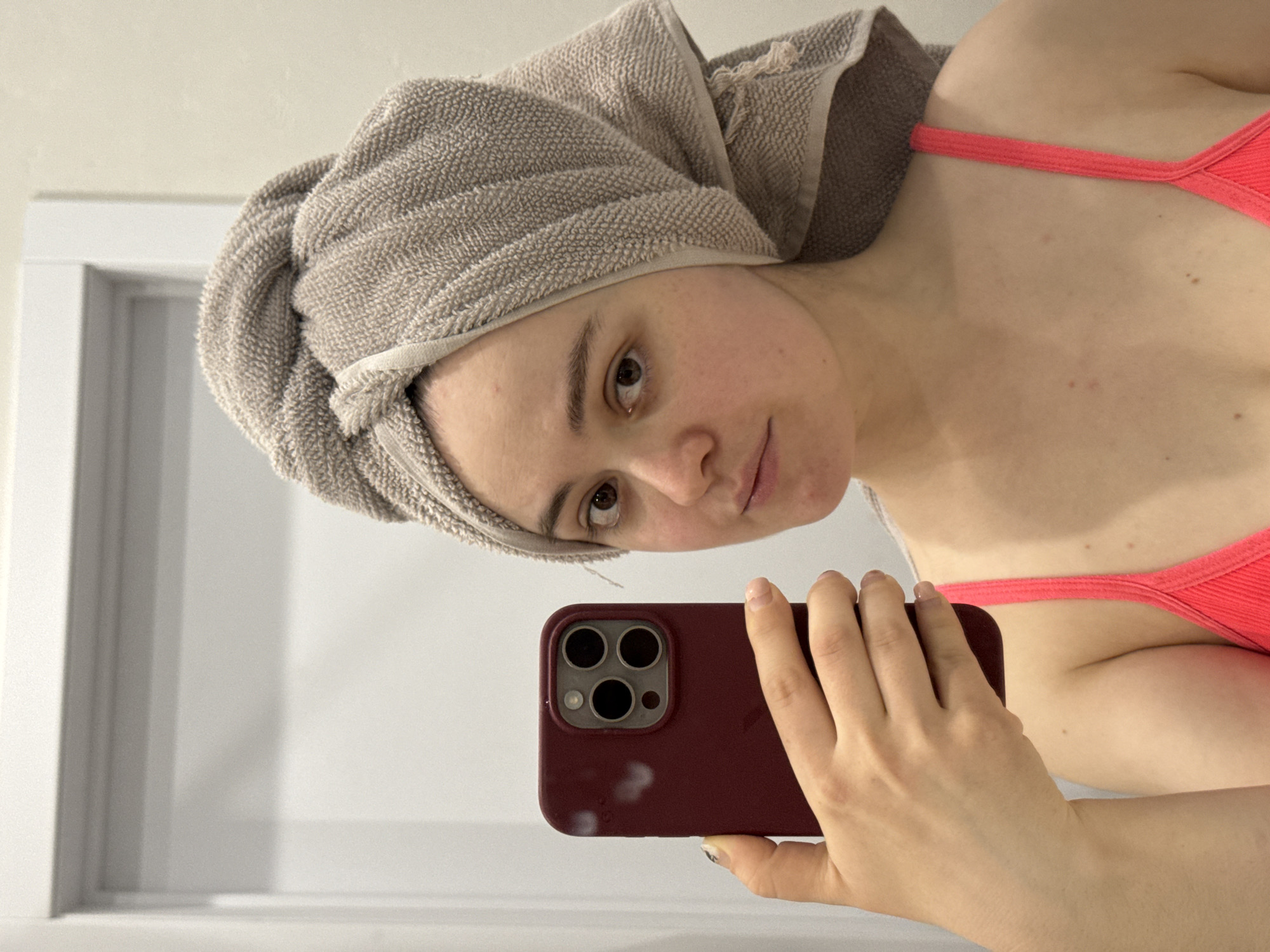 A person is holding a maroon-colored iPhone with three rear cameras, taking a selfie in what appears to be a bathroom. The individual has a towel wrapped around their head and is wearing a red top. The background shows a white doorframe and wall. The location in the city of Izhevsk, Russia, is unknown.