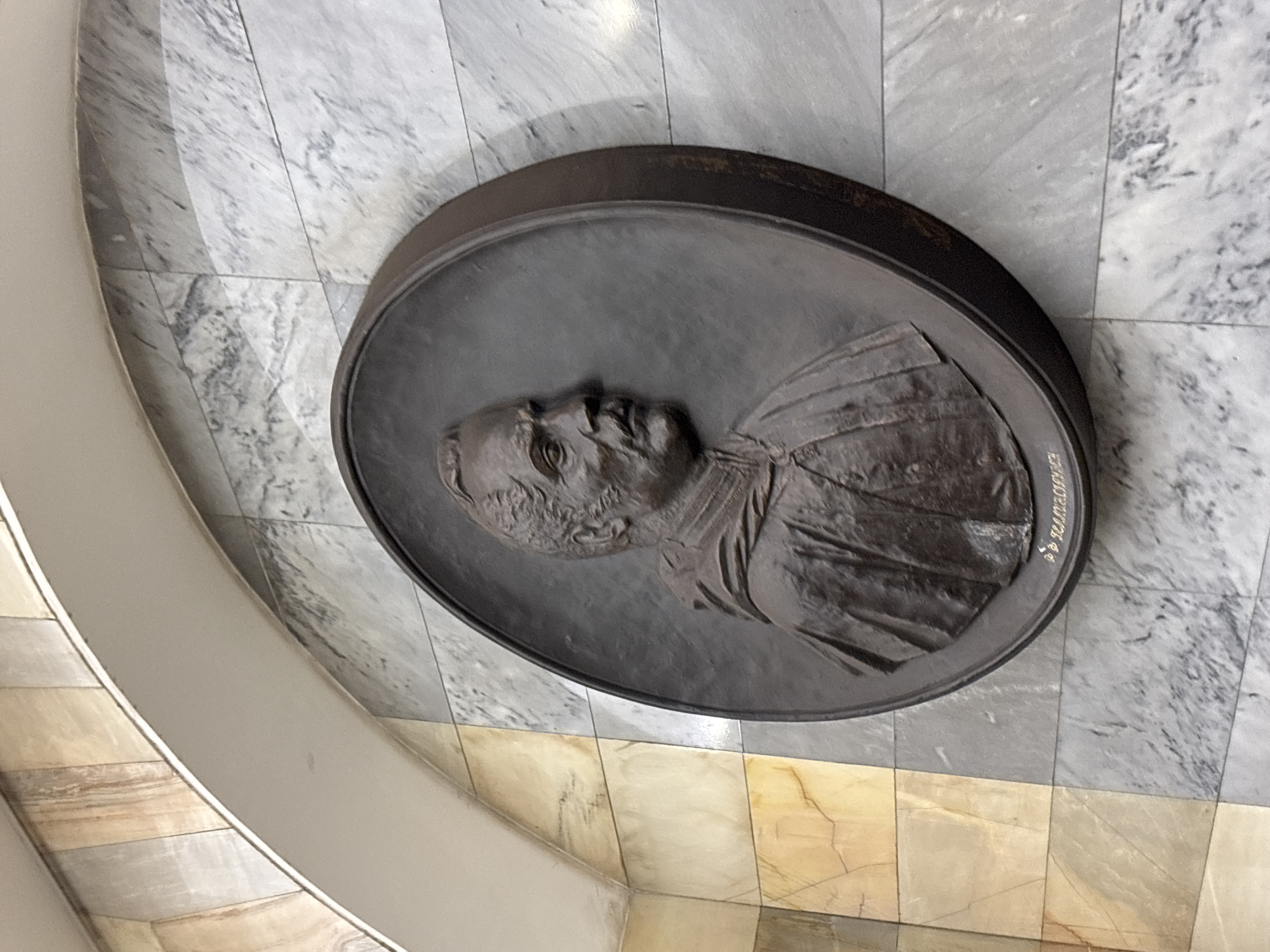 This image shows a bronze relief portrait of someone, mounted on a wall made of light-colored marble tiles. The person in the relief appears to be elderly, with a receding hairline, and is wearing a robe-like garment. The portrait has a signature or inscription on the right side, though the text is not fully legible. The wall on which the relief is mounted is made up of square marble tiles with a mottled pattern. A curved, light-colored wall can be seen along the left side of the image, suggesting this may be a rotunda.
Without additional information, it is impossible to pinpoint the precise location. Given the style of architecture and art, along with the image including Russian text, it is likely located in Saint Petersburg, Russia.