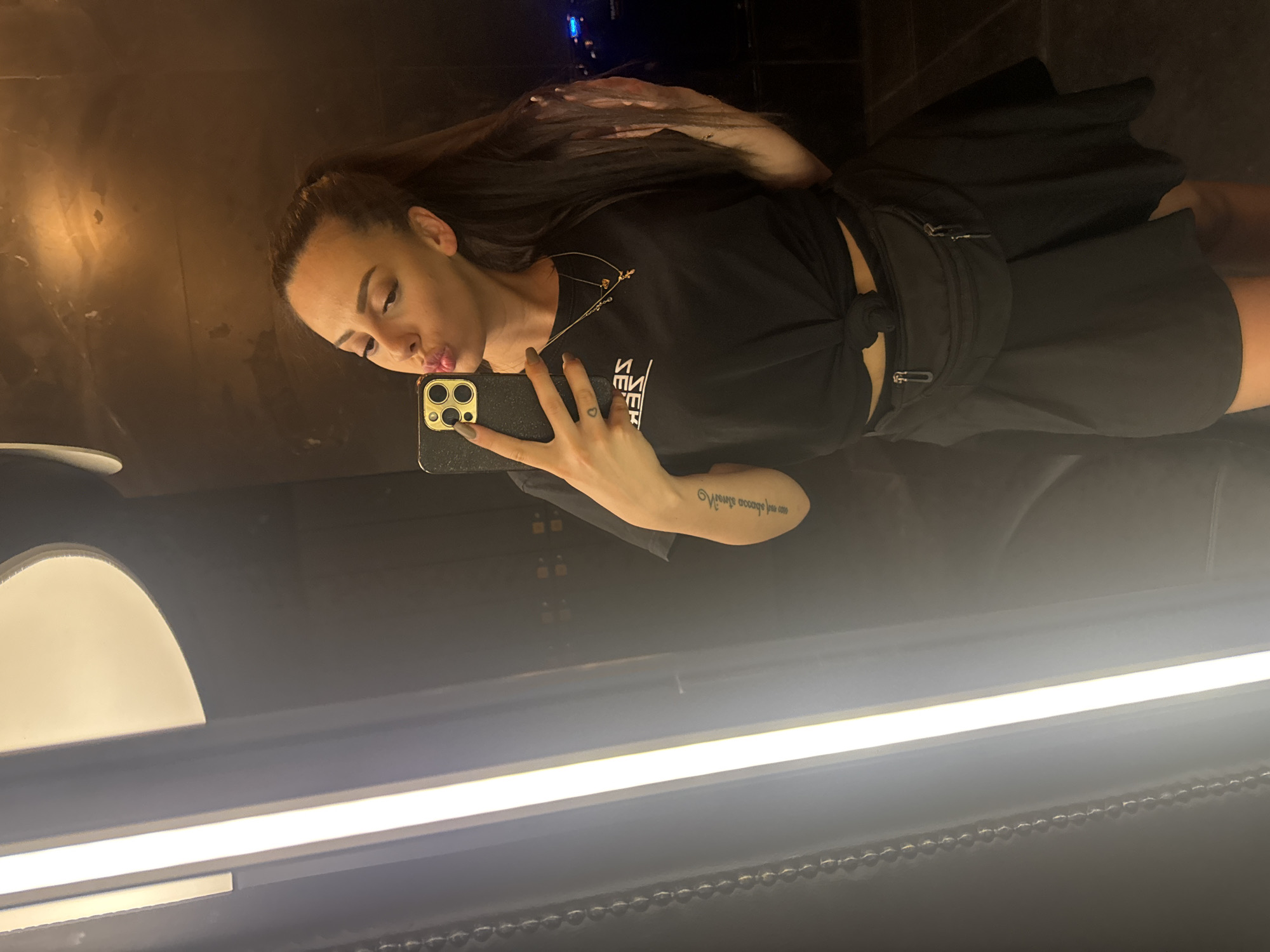 In Varna, Bulgaria, a young woman with dark hair tied in a high ponytail takes a selfie in a dimly lit room with a large, brightly lit vertical panel on the left. She is wearing a black t-shirt tied at the waist, a black fanny pack, and a black skirt. Her left arm is extended, holding a gold-accented smartphone, and her lips are puckered in a kiss. A tattoo is visible on her left forearm. She is also wearing a gold necklace with a small pendant. The background appears to be a reflective surface with some distant lights.