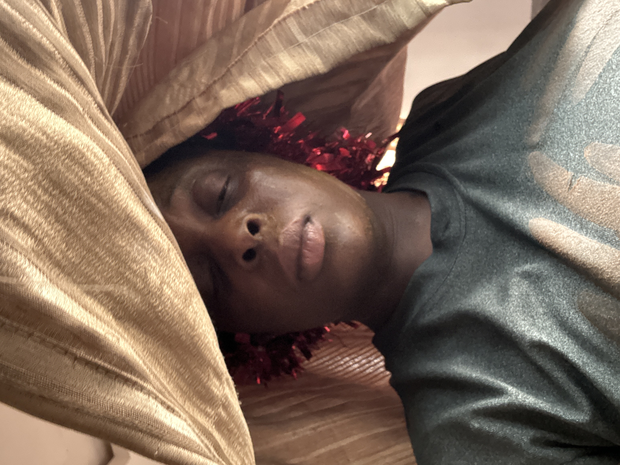 A close-up shot shows a person with dark skin lying down, eyes closed, seemingly asleep. The person is partially covered by a gold-colored, textured fabric that appears to be a pillow or bedding. Red tinsel is draped around their head. They are wearing a dark-colored shirt. The image gives no indication as to the specific location in Sabon Tasha, Nigeria, or even that it was taken there. There are no discernible interactions or actions taking place other than the person lying down.