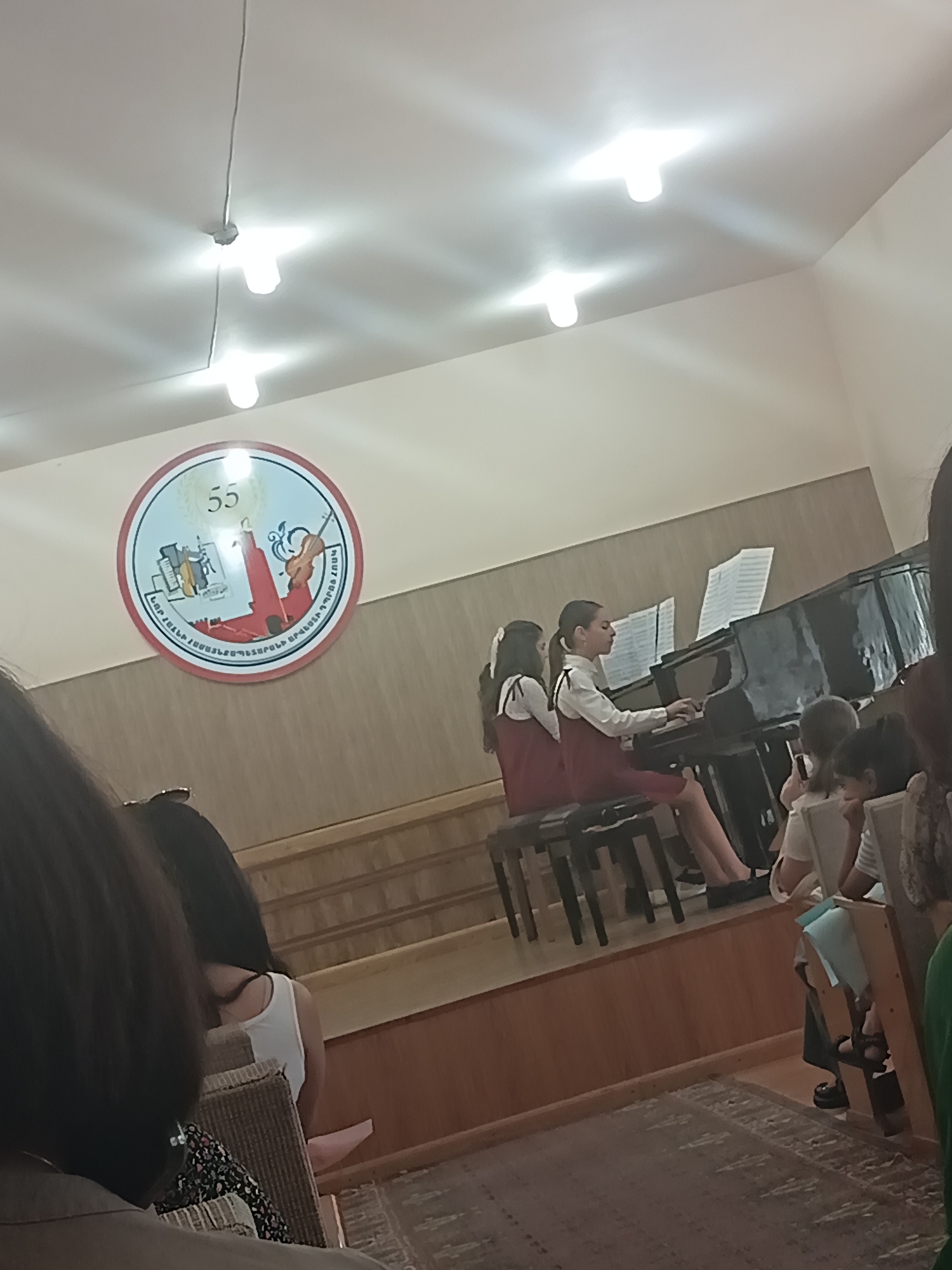 In an indoor setting with a beige ceiling, two young girls are seated on a bench in front of a grand piano, playing a duet. One girl is wearing a white shirt and a maroon dress, while the other is wearing a similar maroon dress over a white shirt. Both have their hair pulled back. Sheet music is visible on the piano. Behind them, a few other individuals, including children and adults, are seated in the audience, with some partially visible in the foreground. The room has wooden paneling on the walls and a carpeted floor. A circular logo with the number "55" and various artistic elements, possibly representing musical instruments, is displayed on the wall above and to the left of the piano. The lighting appears to be from overhead fixtures, casting a bright glow.