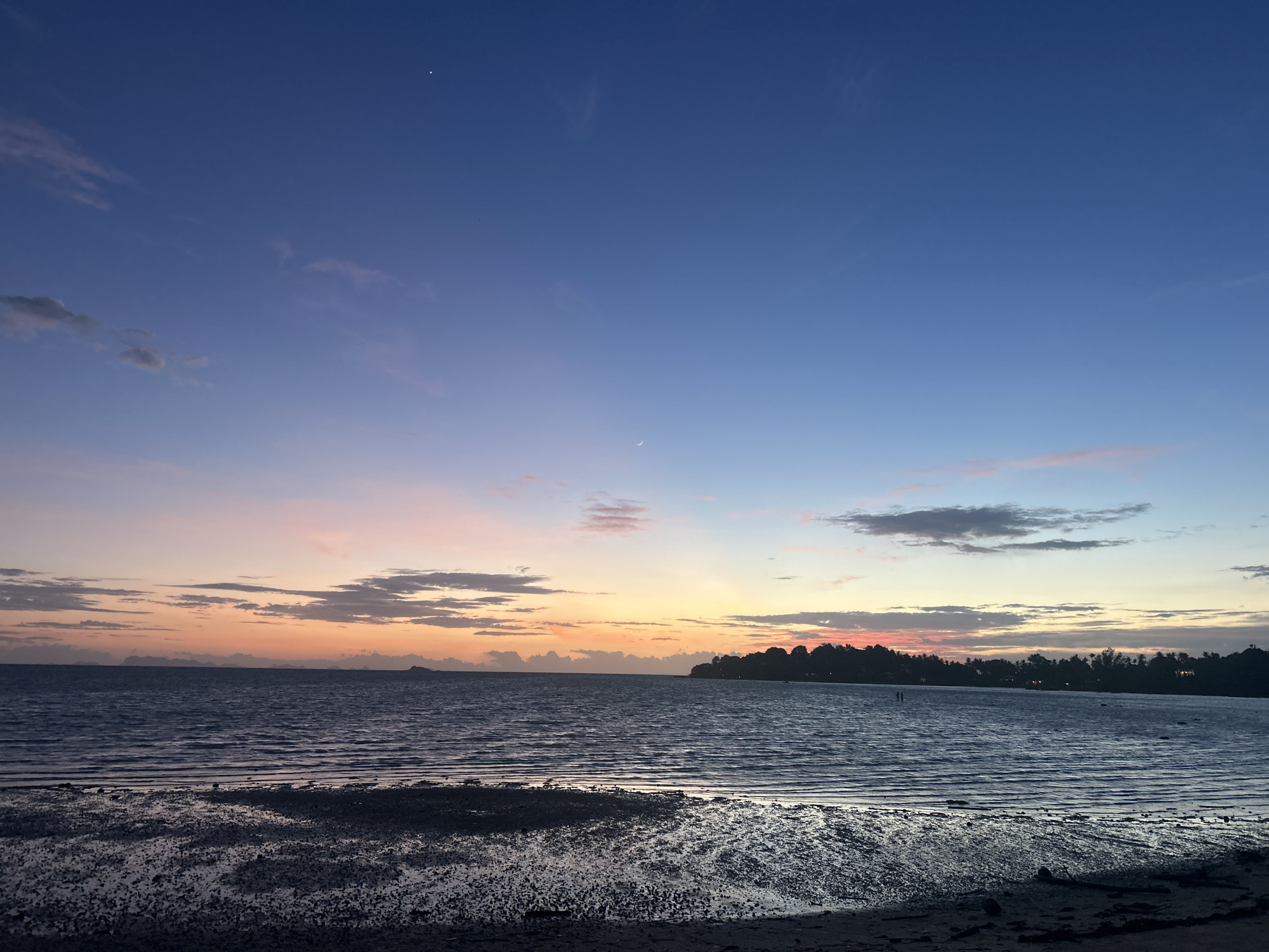 The image depicts a serene coastal scene during twilight in Ko Pha-ngan District, Thailand. The sky transitions from a deep blue at the top to hues of pink, orange, and yellow near the horizon, indicating either sunrise or sunset. Wispy clouds are scattered across the sky, some illuminated by the fading light. A thin crescent moon and a bright star are visible in the upper portion of the sky.
The foreground is a muddy, wet shoreline with reflections on its surface. The water stretches out to the horizon, with gentle ripples on its surface. In the distance, a tree-lined silhouette of the coastline is visible, with hints of human habitation, including what appear to be small structures. Two indistinct figures can be seen standing in the shallow water, suggesting the presence of people enjoying the evening. The overall atmosphere is tranquil and picturesque, characteristic of a tropical coastal evening.