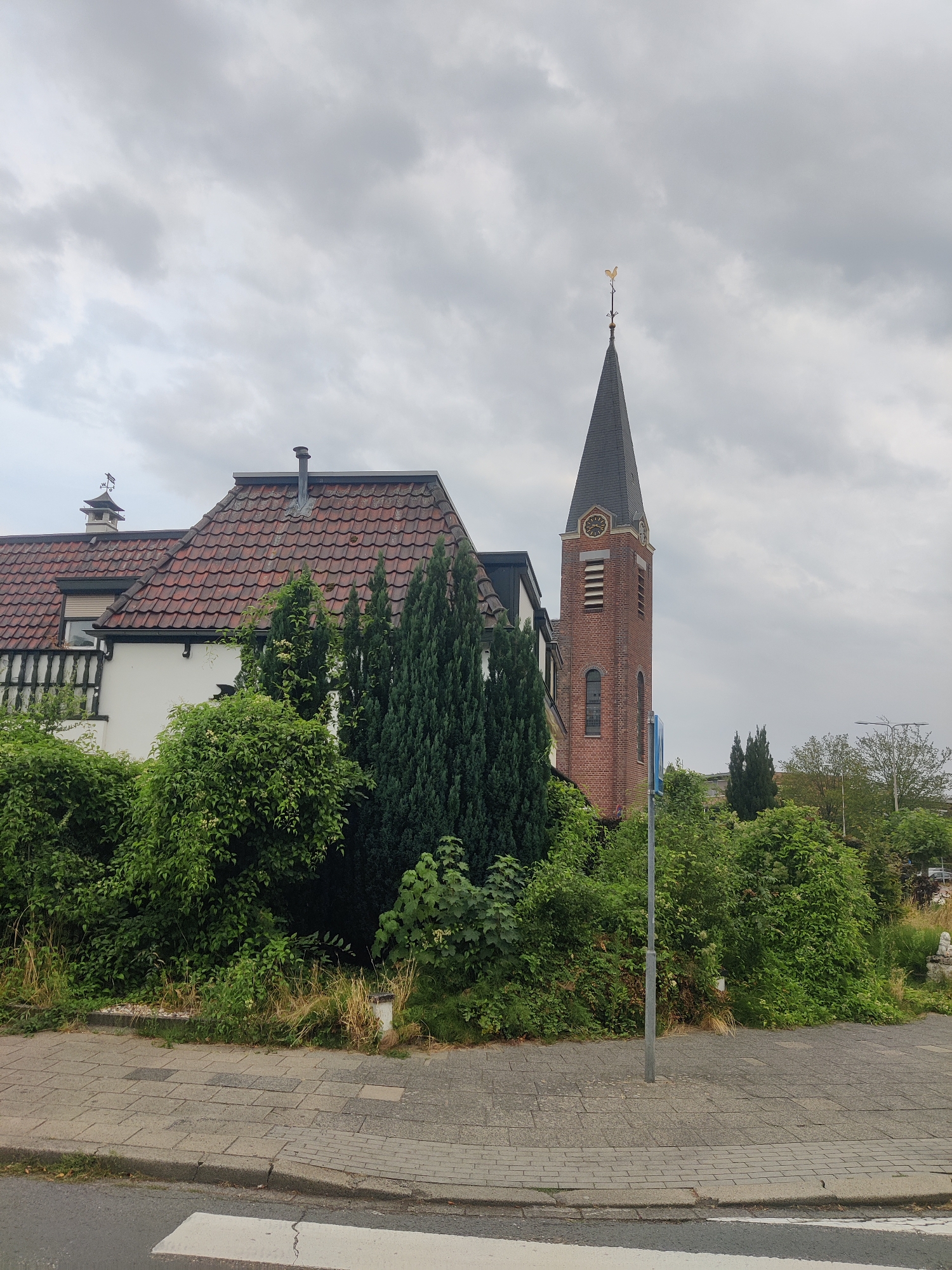 The image shows a brick church tower with a pointed roof and a golden weather vane, located in the city of Winterswijk, Netherlands. To the left of the tower, a house with a red tiled roof and white walls is visible, partially obscured by lush green bushes and trees. A paved sidewalk with a crosswalk in the foreground suggests a public area. The sky is overcast, indicating a cloudy day. No people are present in the image. The scene appears to be a residential area or a part of the city with both residential and religious buildings.
