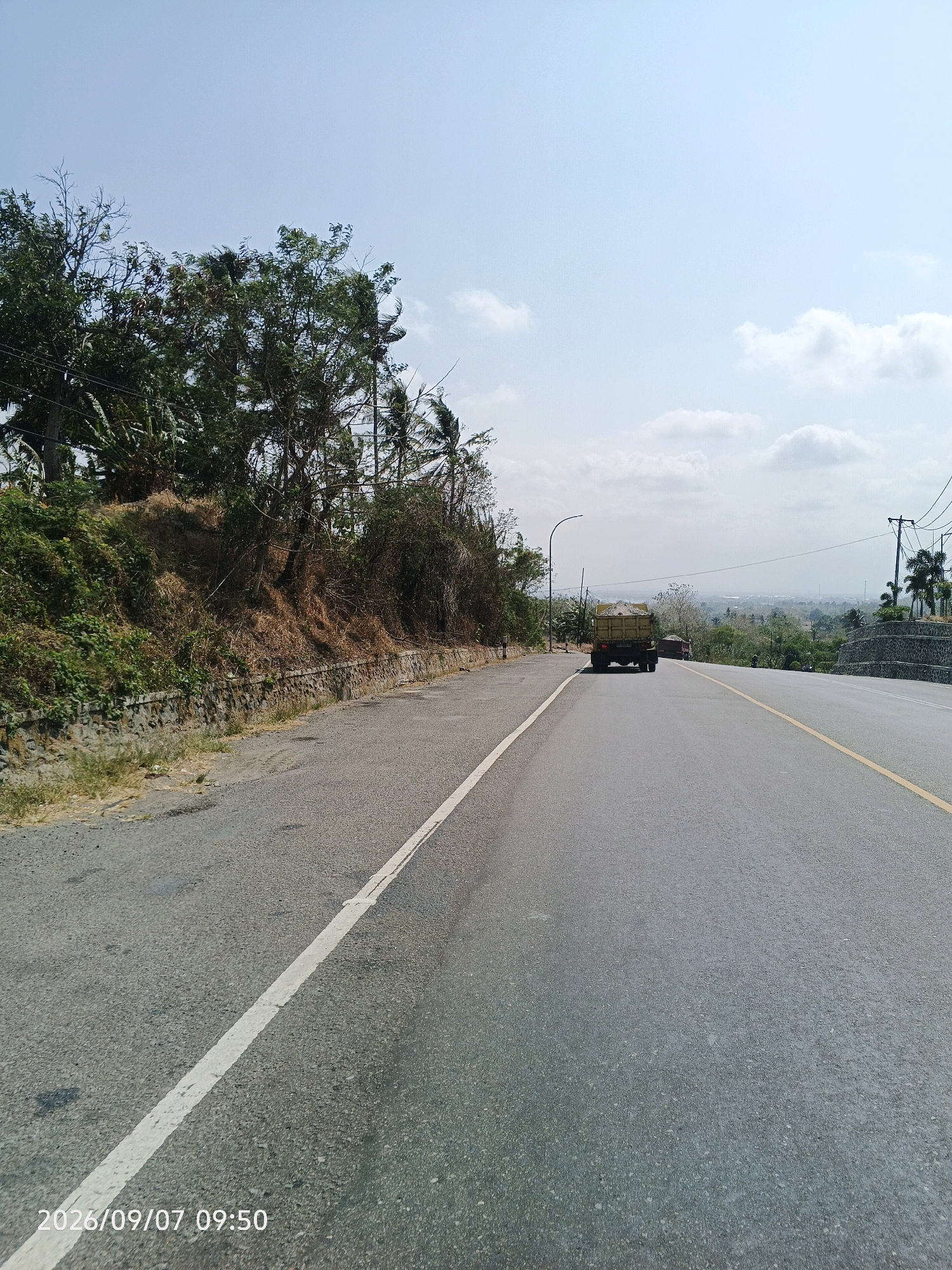 A wide asphalt road with a white line marking the left edge and a yellow line marking the center stretches into the distance under a clear blue sky with scattered white clouds. On the left side of the road, a steep embankment is covered with green vegetation and dry brown grass, with a low stone wall at its base. Trees and foliage grow along the top of the embankment. In the distance, a yellow dump truck with a load of material is driving away from the camera, followed by another vehicle. The landscape beyond the road features rolling hills and a hazy view of a town or city. Utility poles with wires are visible on the right side of the road. The timestamp "2026/09/07 09:50" is visible in the lower left corner of the image. The scene appears to be in Lawawoi, Indonesia, given the typical road and vegetation seen in the region.