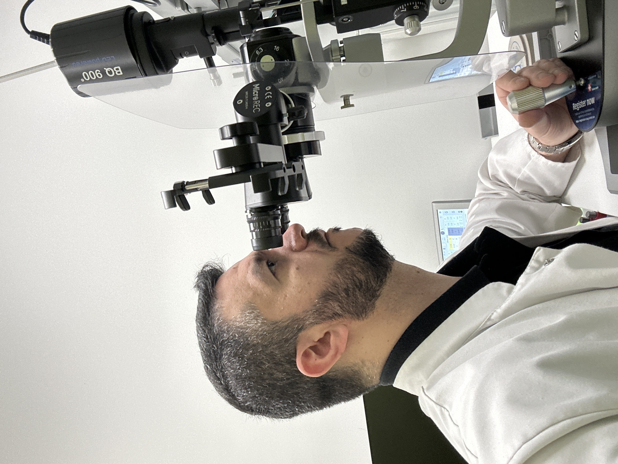 An adult male with dark, greying hair and a beard is undergoing an eye examination in what appears to be a medical or optometry clinic in Berlin, Germany. He is seen in profile, looking upwards into an ophthalmic examination machine, likely a slit lamp. His right ear is visible.
The examination equipment is prominent, featuring black cylindrical components, various silver knobs, and a clear plastic shield. One black component bears the text "LED powered" and "BQ 900°". Another section of the device shows "OCE O" and "MicroREC", along with "MAGE 12054", "6.3", and "I". An unseen individual, presumably the examiner, operates the machine with a hand visible holding a silver-colored textured knob. This hand wears a silver watch with a metal band. The examiner is wearing a white lab coat or medical uniform.
In the background, a digital screen with graphic data is partially visible. A small label with the text "Register now" is also attached to the machine. The setting is bright with predominantly white walls or surfaces. The overall activity depicts a routine or specialized eye care procedure.