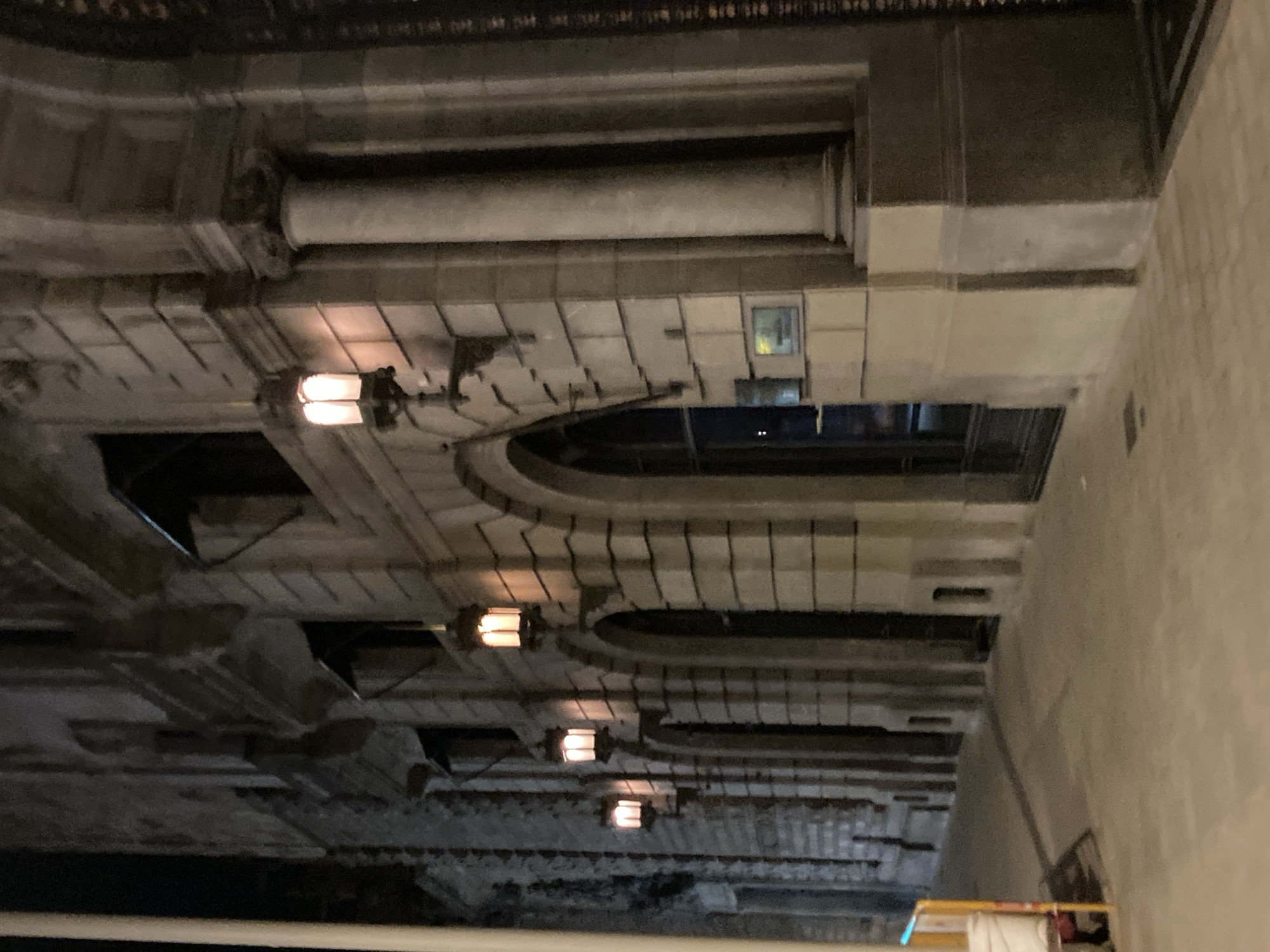 This is a low-angle, nighttime exterior shot of a building in Barcelona, Spain. The building facade is constructed of stone blocks, with arched doorways and windows. Several ornate lanterns, emitting a warm, yellow light, are mounted on the building's exterior at intervals along the facade. The pavement in front of the building is illuminated by the lanterns, casting shadows. A tall, thin white pole is visible on the far left of the frame, likely a street lamp or utility pole. To the far left, a yellow and white barrier is partially visible, suggesting ongoing construction or a temporary obstruction. The overall impression is one of an older, possibly historic, urban environment at night.