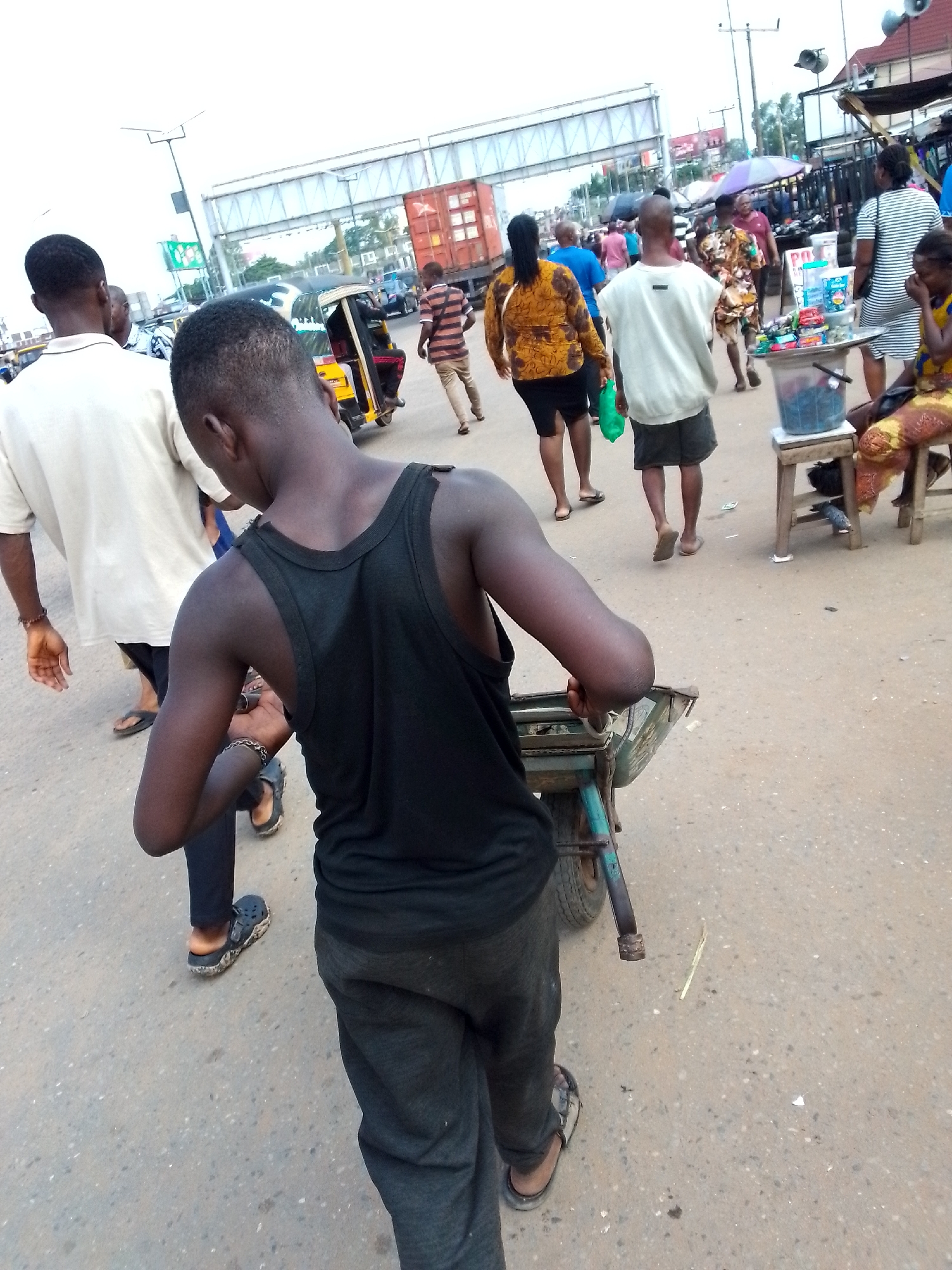 This is a street-level photograph taken in Onitsha, Nigeria. In the foreground, a young Black male wearing a black tank top and dark pants is walking away from the camera, pushing a metal cart with two wheels. He has a bracelet on his left wrist. To his left and slightly behind him, a Black male in a white t-shirt and black shorts walks away from the camera. Further back, a group of people are visible walking in the street. These include a person in a striped shirt and khaki pants, a woman in a yellow and orange patterned dress, and a person in a white shirt and dark shorts. In the background, there are market stalls, a yellow and black auto-rickshaw, and a large overhead structure that appears to be part of a bridge or overpass. A red shipping container is visible on a truck. The ground is asphalt, and there are various scattered items and debris on the street. The sky is overcast.