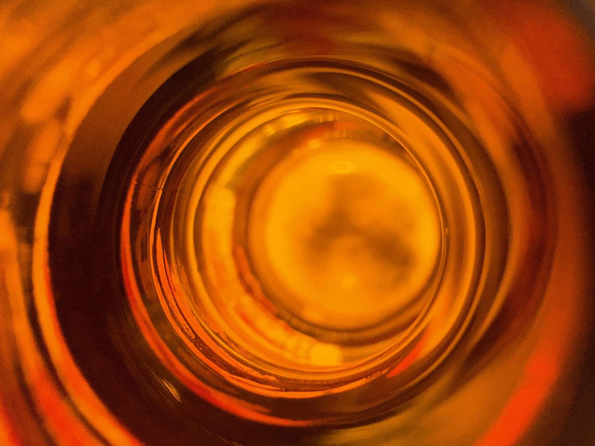 The image is a close-up, top-down view into the opening of a dark orange, translucent bottle. Concentric rings of light and shadow create a tunnel-like effect, with the brightest light emanating from the bottom of the bottle. The texture of the glass is visible as subtle ridges and variations in the light. There are no people, animals, or other objects in the image, and no indication of the location being in Novorossiysk, Russia.