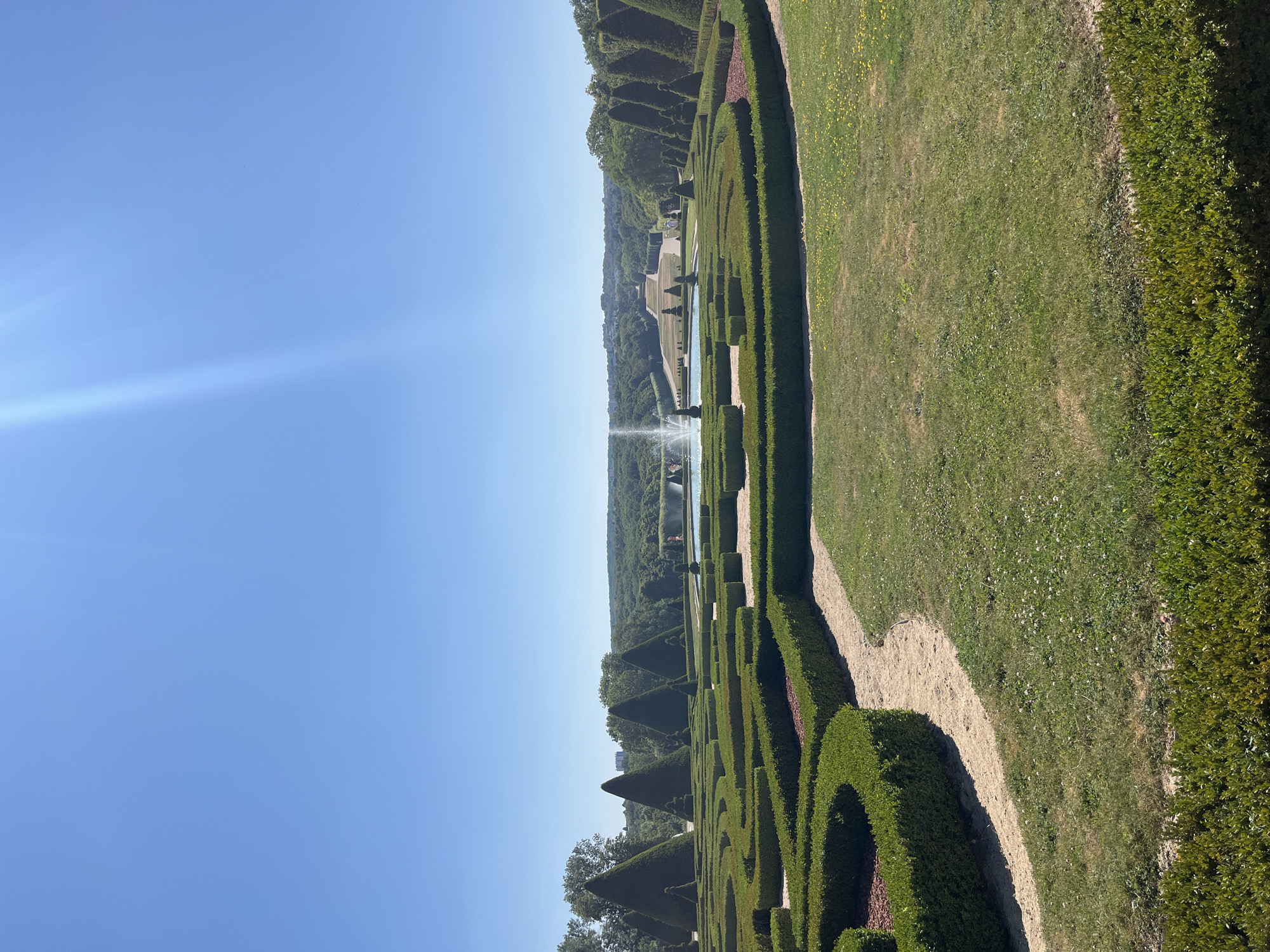 This image shows a view of the formal gardens at the Parc de Sceaux, France, on a clear, sunny day. The foreground features meticulously maintained hedges forming intricate geometric patterns, with a sandy pathway winding through them. Beyond the hedges, a large fountain shoots water into the air in a pool of turquoise water. In the background, conical topiaries line the sides of the garden. Further back, a long, symmetrical pathway stretches towards a distant building, flanked by rows of trees and rolling, verdant hills that form the horizon. The sky is a bright, cloudless blue, with a subtle lens flare in the upper right quadrant. There are no visible people in the immediate foreground, but there are a few small figures visible further back in the garden near the fountain and pathway. The overall impression is one of grand, manicured landscape design.