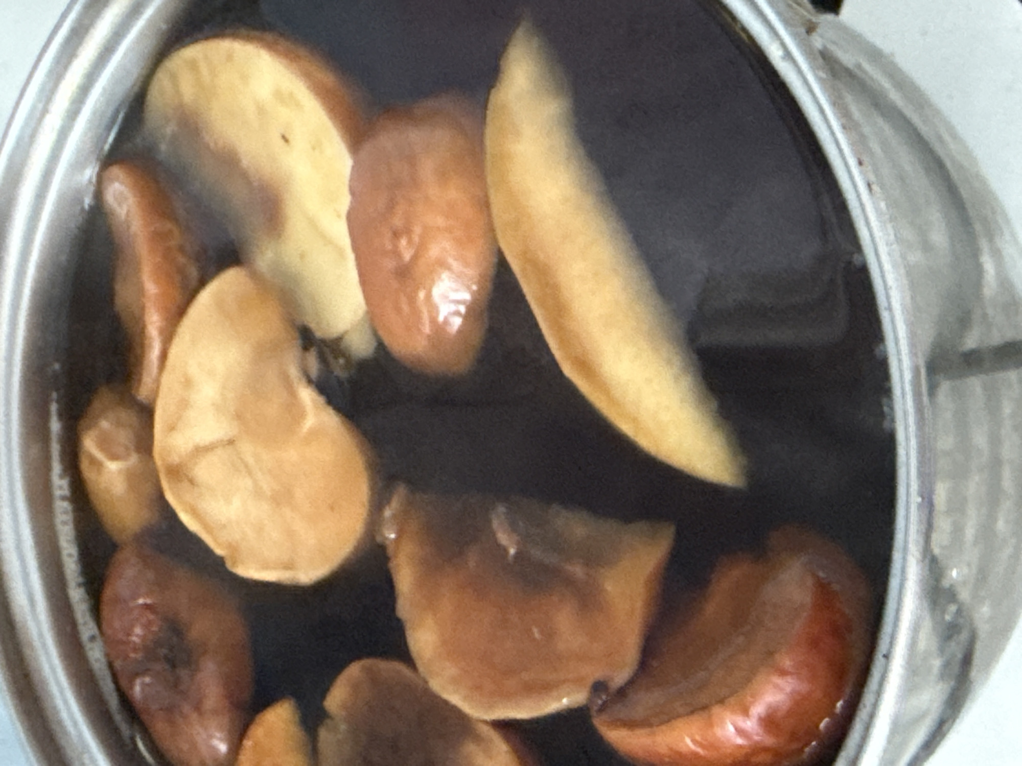 A close-up, overhead shot shows pieces of what appear to be apples or pears simmering in a dark liquid within a metal pot. The fruit slices are a warm, golden-brown and some have a reddish hue, suggesting they may be caramelized or a variety with colored skin. The liquid is dark, indicating a rich broth or syrup. The pot's rim is visible, along with some reflections on its metallic surface. The overall impression is of food being prepared, possibly for a dessert or a warm beverage. The scene is devoid of any specific geographical markers, making it impossible to determine a location within a city.