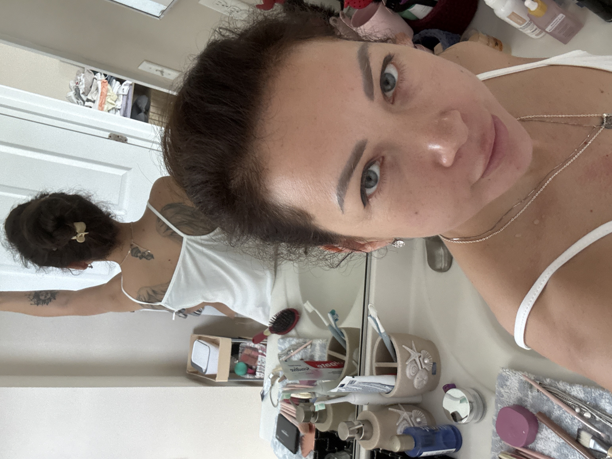 This media file shows a woman standing in what appears to be a bathroom. She is reflected in a mirror, which allows for a view of both her front and back. The woman has dark hair styled in a bun with a gold clip. She is wearing a white tank top and has several tattoos on her back and arm. In the foreground, the woman is looking towards the camera, her face close to the lens. She has blue eyes, dark eyebrows, and pink lipstick. A silver necklace with a pendant is visible around her neck.
The bathroom counter is cluttered with various items, including a toothbrush holder with toothbrushes and toothpaste, a hairbrush, a small box with a white object inside, and several bottles and containers of toiletries. A light-colored towel is also on the counter, with makeup brushes and a purple object on top of it. The overall setting suggests a personal grooming area. The lighting in the image is bright, suggesting it might be daytime, although no specific weather or time of day cues are definitive. The reflection in the mirror also shows a closet or shelf with clothing items. The woman's expression is neutral, and she is taking a selfie. No discernible text is visible, except for what appears to be branding on a toothpaste tube which reads "Colgate".