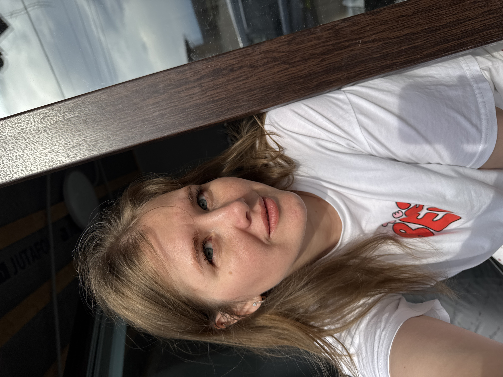 A woman with blonde hair and blue eyes is lying down, wearing a white t-shirt with a red design. She appears to be indoors near a window with a dark brown frame. Reflected in the window are exterior buildings and sky. In the lower left corner, the word "JUTAPO" is visible in reverse. The location is inferred to be in Gornyi (Dvurechenskoe S/P), Russia.