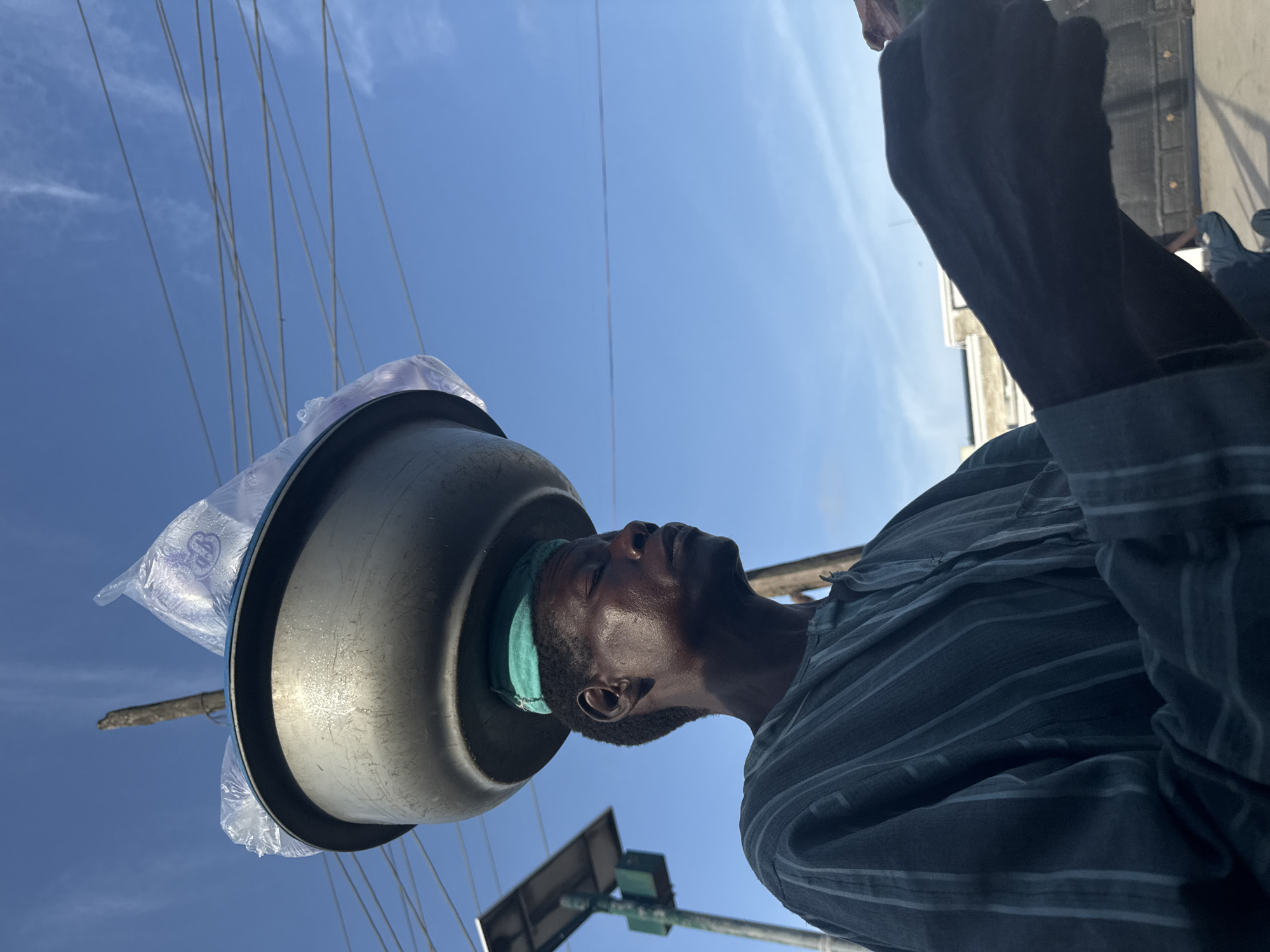 A dark-skinned adult male is depicted in a profile view facing right, illuminated by bright daylight against a clear blue sky, in the city of Ado, Nigeria. He wears a dark, vertically striped, long-sleeved shirt. A green textile pad is positioned on his head, cushioning a large, scratched, dark-grey metallic basin. The basin contains several transparent plastic bags, one of which exhibits a purple graphic. His eyes are downcast or closed. His visible right hand, positioned in the lower right foreground, holds a small, dark, rectangular object. In the left mid-ground, a slender pole supports a small solar panel and an attached green utility box. Multiple thin, light-colored electrical wires traverse the upper half of the sky. Distant building outlines and a dark, possibly wooden, vertical post are present in the background.