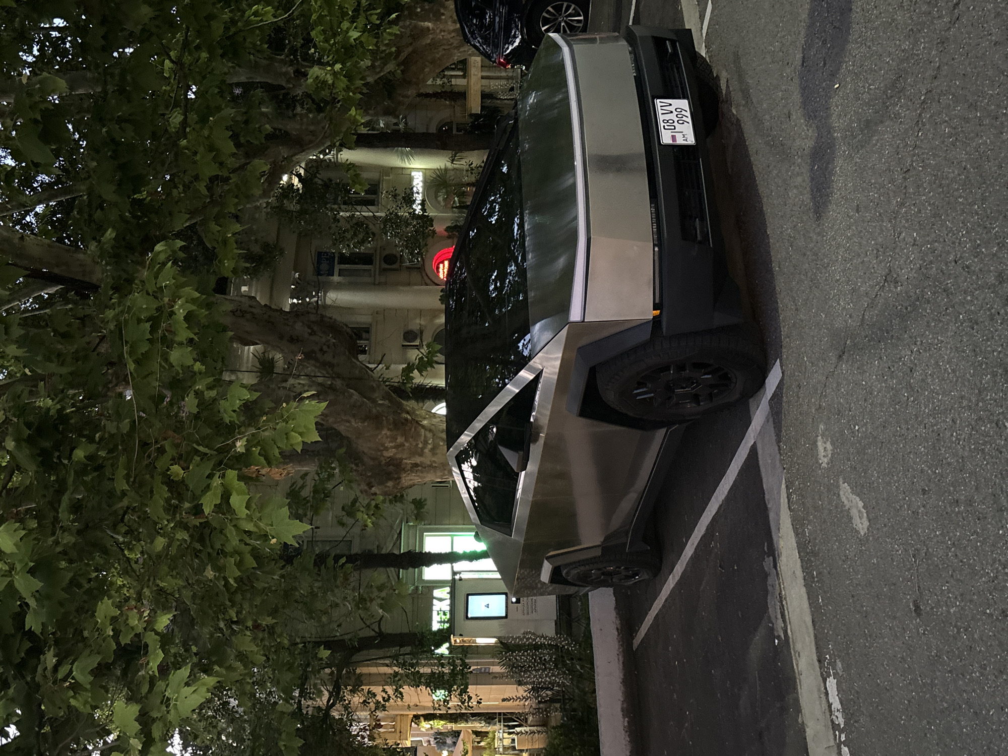 The image shows a Tesla Cybertruck parked on an asphalt surface, likely a street or parking lot, in Sochi, Russia. The vehicle is the primary subject, with its distinctive angular, metallic design clearly visible. It appears to be nighttime or dusk, given the ambient lighting conditions and the artificial lights illuminating the background buildings.
The setting includes a tree with lush green foliage in the foreground, partially obscuring the view of the buildings behind it. The buildings in the background appear to be multi-story structures with windows and architectural details typical of an urban environment. One building has a red traffic light visible.
There are no people clearly visible in the image. The Cybertruck has a license plate that reads "GB VV AM 999". The overall mood is quiet and still, perhaps indicating a moment of rest or display of the futuristic vehicle in its environment. The weather appears calm, with no indication of rain or strong winds. The presence of streetlights and building lights suggests it is an urban area within Sochi.