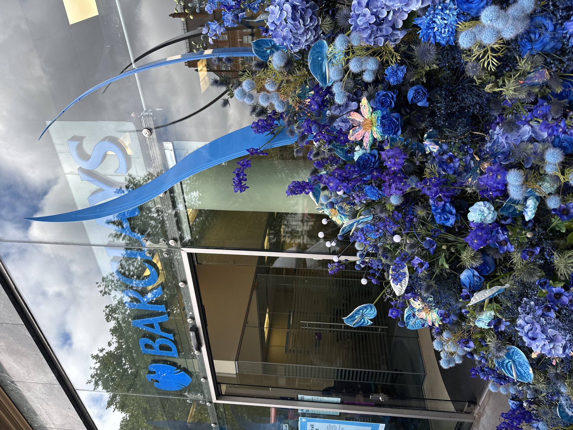The image shows the entrance to a Barclays bank branch in London, United Kingdom. The bank's logo is visible on the glass facade. The entrance is decorated with an elaborate floral arrangement in various shades of blue, including hydrangeas, roses, thistles, and anthuriums, as well as decorative elements such as iridescent butterflies and small white orbs. The reflection in the glass shows the sky with clouds and the opposite side of the street with trees. Inside the bank, there are sliding glass doors and signage.