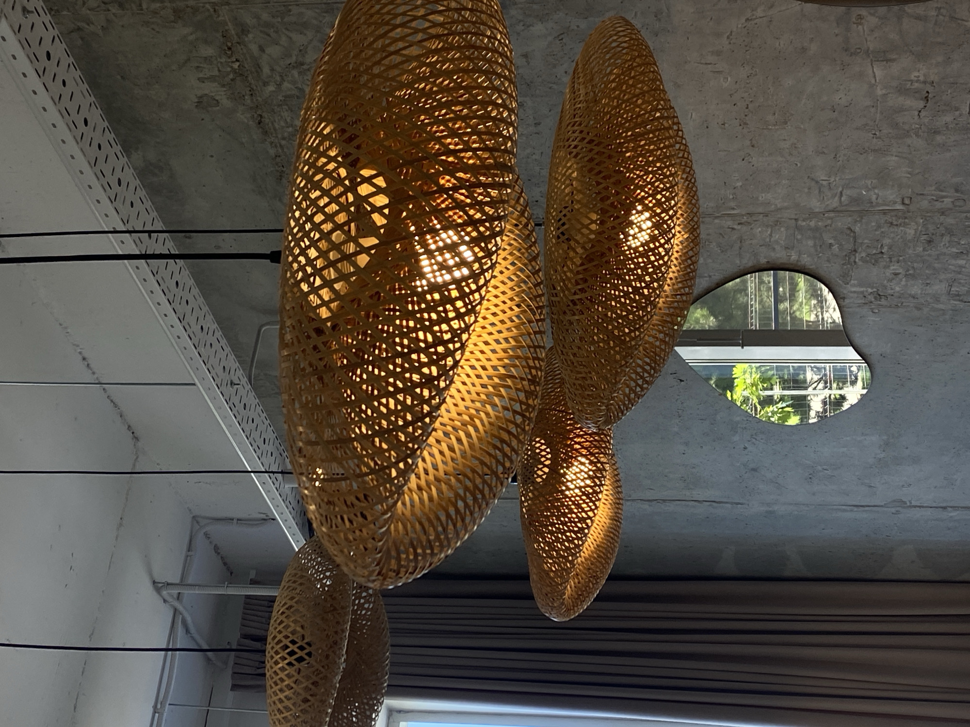 The image depicts a close-up, interior shot of several woven bamboo pendant lights hanging from a ceiling. The lights are an earthy, golden-brown color and have an intricate, open weave pattern that allows the warm light from the bulbs to filter through, creating a soft glow and casting shadows. The ceiling is a combination of white and concrete textures, with visible electrical conduits and fixtures. One of the lights is particularly large and dominant in the foreground, while others are smaller and positioned behind it, creating a sense of depth. To the left, a muted brown curtain is visible, suggesting a window or doorway. In the lower right portion of the image, a round, abstractly shaped mirror is attached to a textured concrete wall. The mirror reflects an outdoor scene with green foliage and what appears to be a window frame. The overall aesthetic suggests a modern, yet natural and cozy ambiance. The location is a room in the city of Dinskaya, Russia.