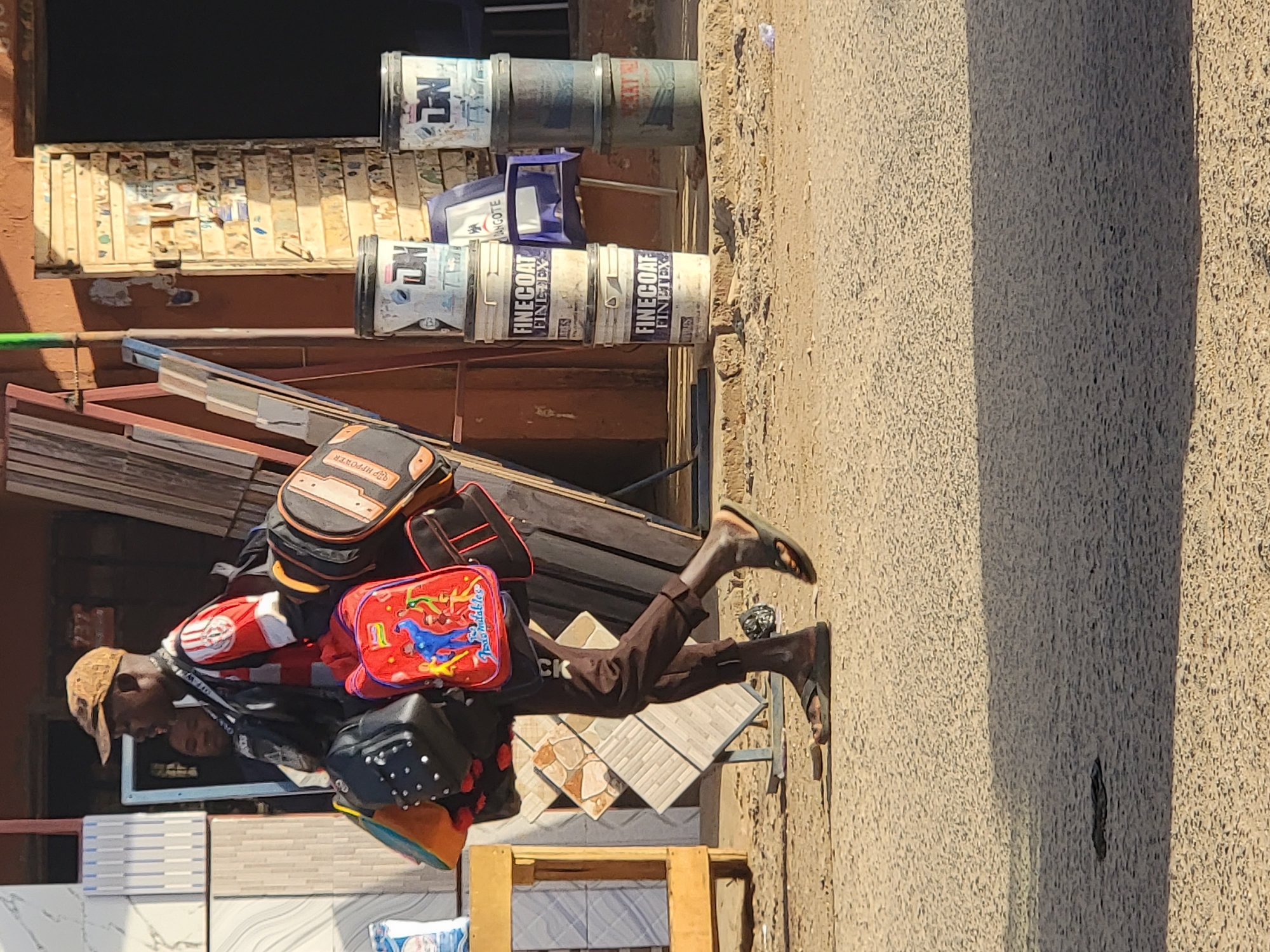 A man is walking on a sidewalk in Maiburiji, Nigeria. He is carrying several bags and wearing a cap and sandals. Various objects are situated behind the man. These include paint cans, tile displays, and construction materials.