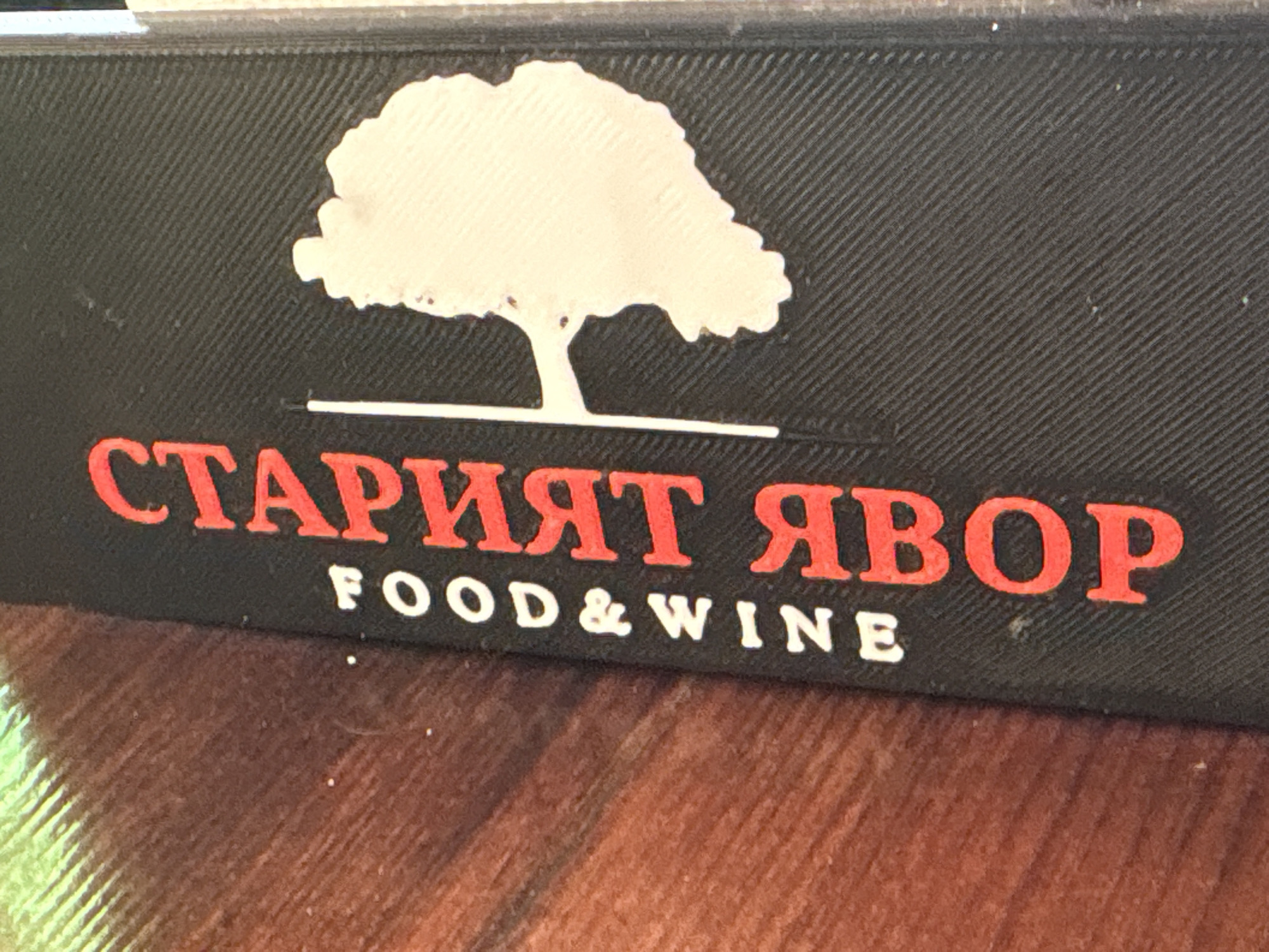 The image shows a close-up of a black surface with a textured pattern. A white silhouette of a tree with a thick trunk is centered at the top of the surface. Below the tree, in large red Cyrillic letters, are the words "СТАРИЯТ ЯВОР". Beneath these red letters, in smaller white Latin letters, are the words "FOOD & WINE". The surface appears to be part of a sign or packaging. The background is a blurred wooden surface with warm, reddish-brown tones and visible wood grain. The lighting creates some glare on the surface. The location is identified as Parvenets, Bulgaria.