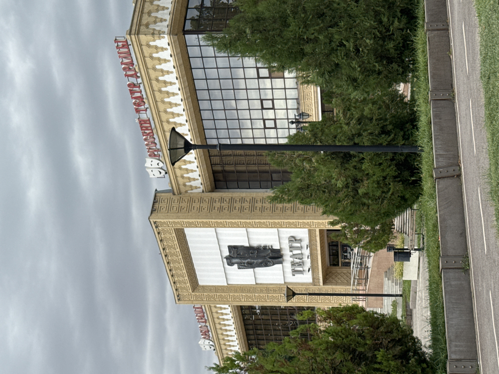 The image shows the Russian Drama Theatre in Shymkent, Kazakhstan. The building features a prominent facade with decorative elements and signage. On the left side of the building, there is a large relief sculpture set within a rectangular panel. Below this sculpture, the word "TEATP" is displayed in large, capitalized letters. Above this, there is a smaller sign with what appears to be Cyrillic text. On the roofline, there are two signs with red Cyrillic lettering, one on the left reading "РУССКИЙ ТЕАТР" and another on the right that is partially obscured but appears to continue the phrase. Two theatrical masks, one smiling and one frowning, are also visible near the left roofline sign. The building has large glass windows that reflect the cloudy sky. In front of the building, there are several evergreen trees. A tall lamppost stands to the left of the main entrance area, which features stairs and ramps. A bench and a trash can are visible to the right, further along the facade. The foreground shows a road with white lane markings and a concrete barrier. The overall impression is of an overcast day, with the architecture of the theatre being the central focus.
