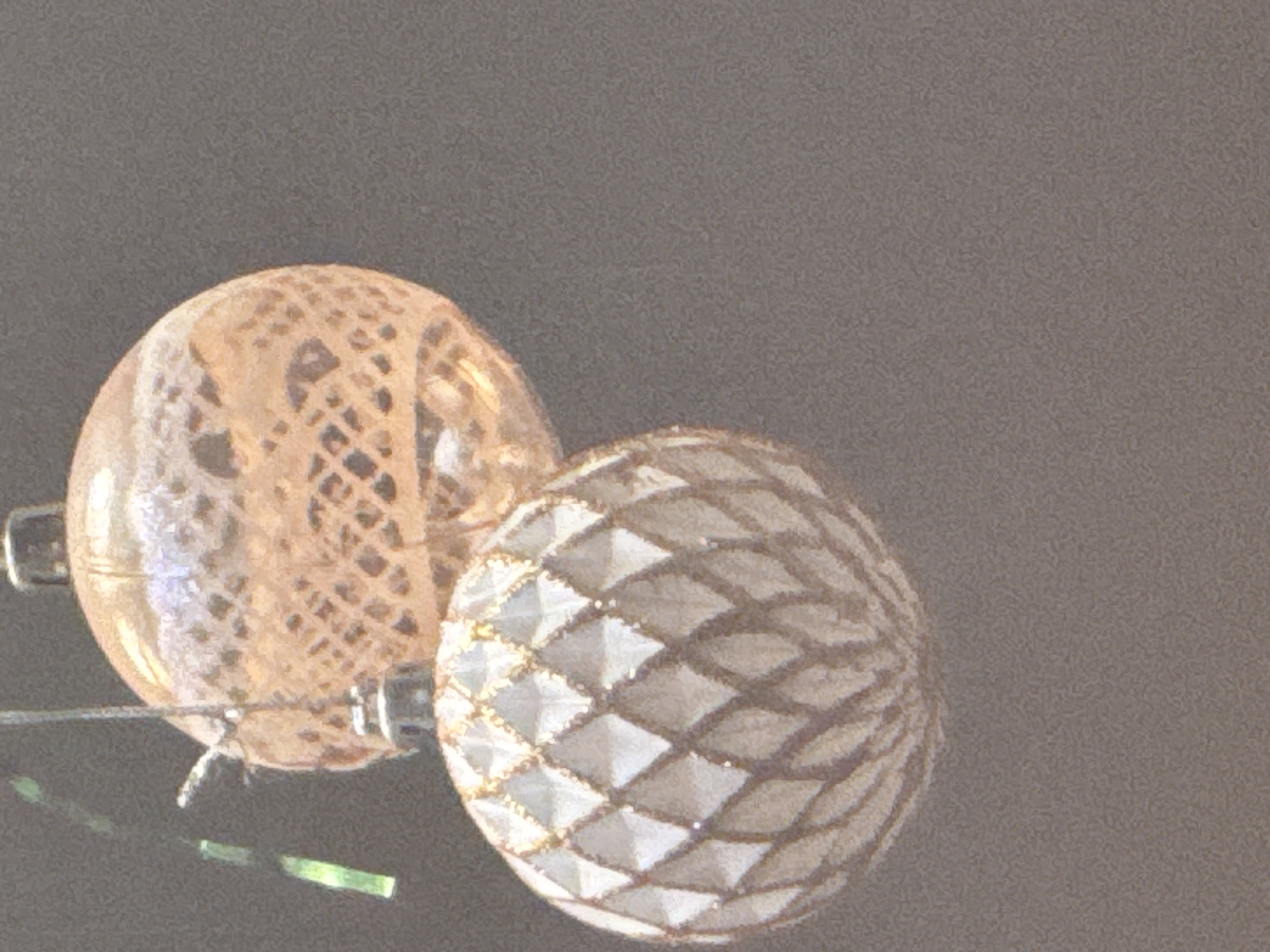 The image shows two ornate, round decorative objects, likely Christmas ornaments, against a muted grey background. The object on the left is translucent and has an intricate, patterned design resembling lace or net, in a warm, orangey-gold hue. The object on the right is opaque and covered in a pattern of raised diamond shapes, outlined in a darker color and filled with a light, possibly silver or white material. Some of these diamond facets catch the light, suggesting a subtle sparkle. Both ornaments appear to be suspended by thin wires or threads, with a slender, reflective surface visible at the bottom, possibly a table or shelf. Small, rectangular green reflections are also visible on this surface, hinting at some ambient light source. The overall impression is one of festive or decorative items, likely indoors given the lighting and setting. No people are visible, and the activity appears to be stationary display. The time of day and weather are not discernible. There is no visible text in the image. The location context of Kholmsk, Russia, does not provide any visual cues within the image itself.