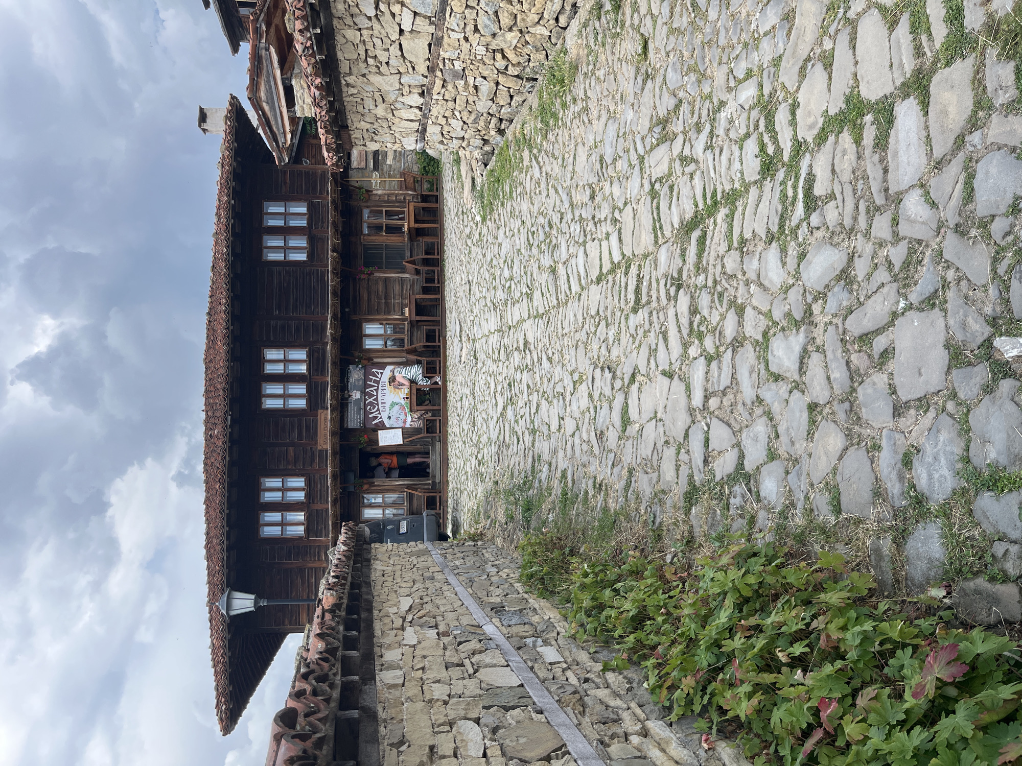 The image shows a cobblestone street leading towards a traditional wooden building in the city of Zheravna, Bulgaria. The building, likely a mehana (tavern), has a dark wood facade with multiple windows and a red tile roof. A sign is visible on the front of the building, featuring Cyrillic script. Stone walls flank the street on either side. There are several tables and chairs set up outside the building, suggesting an outdoor dining area. A few people are visible near the entrance of the building and seated at one of the tables. The sky is overcast, and there is some greenery growing along the stone walls and between the cobblestones.