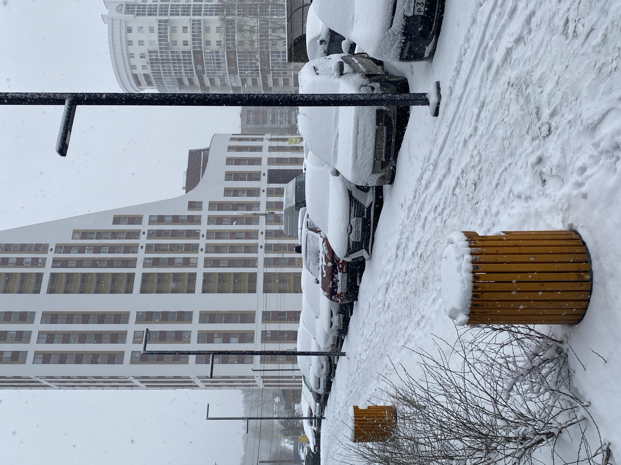 A winter scene in Yekaterinburg, Russia. Cars parked along a snow-covered street are covered in snow. Two modern apartment buildings dominate the background. A wooden bench sits on the sidewalk, also covered in snow, next to a bare tree. A black metal pole, possibly a light post, stands in the foreground. Snow is falling.