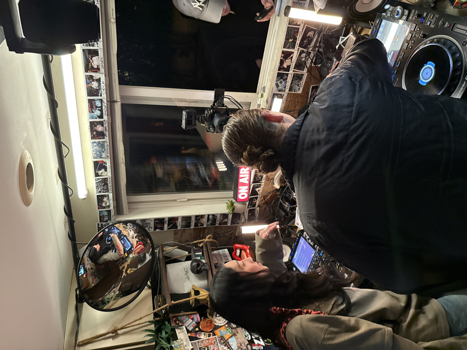 Inside a room, two people are present; one stands wearing a black jacket and the other sits. A DJ mixer is visible near the standing individual. A window is adorned with photos, and an "ON AIR" sign is lit. A camera is mounted near the window. Various stickers cover the lower portion of the room. A convex mirror is mounted on the wall.