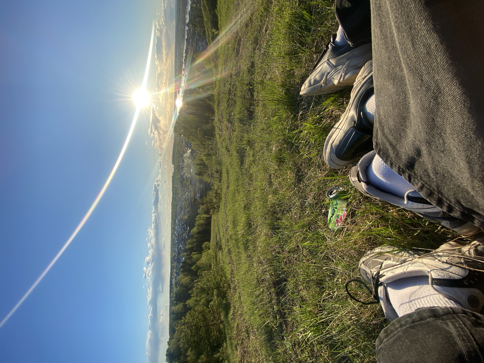 The image depicts a scene on a grassy slope overlooking a city, likely Ольховка, Russia, given the presence of trees, buildings, and a body of water in the distance. The sun is brightly shining, creating a lens flare effect in the sky. In the foreground, three pairs of legs wearing jeans and sneakers are visible. The individuals are sitting on the grass, with their feet pointing towards the viewer. A discarded, crushed can is visible in the grass among the shoes. The overall impression is one of casual relaxation and observation of the landscape.
