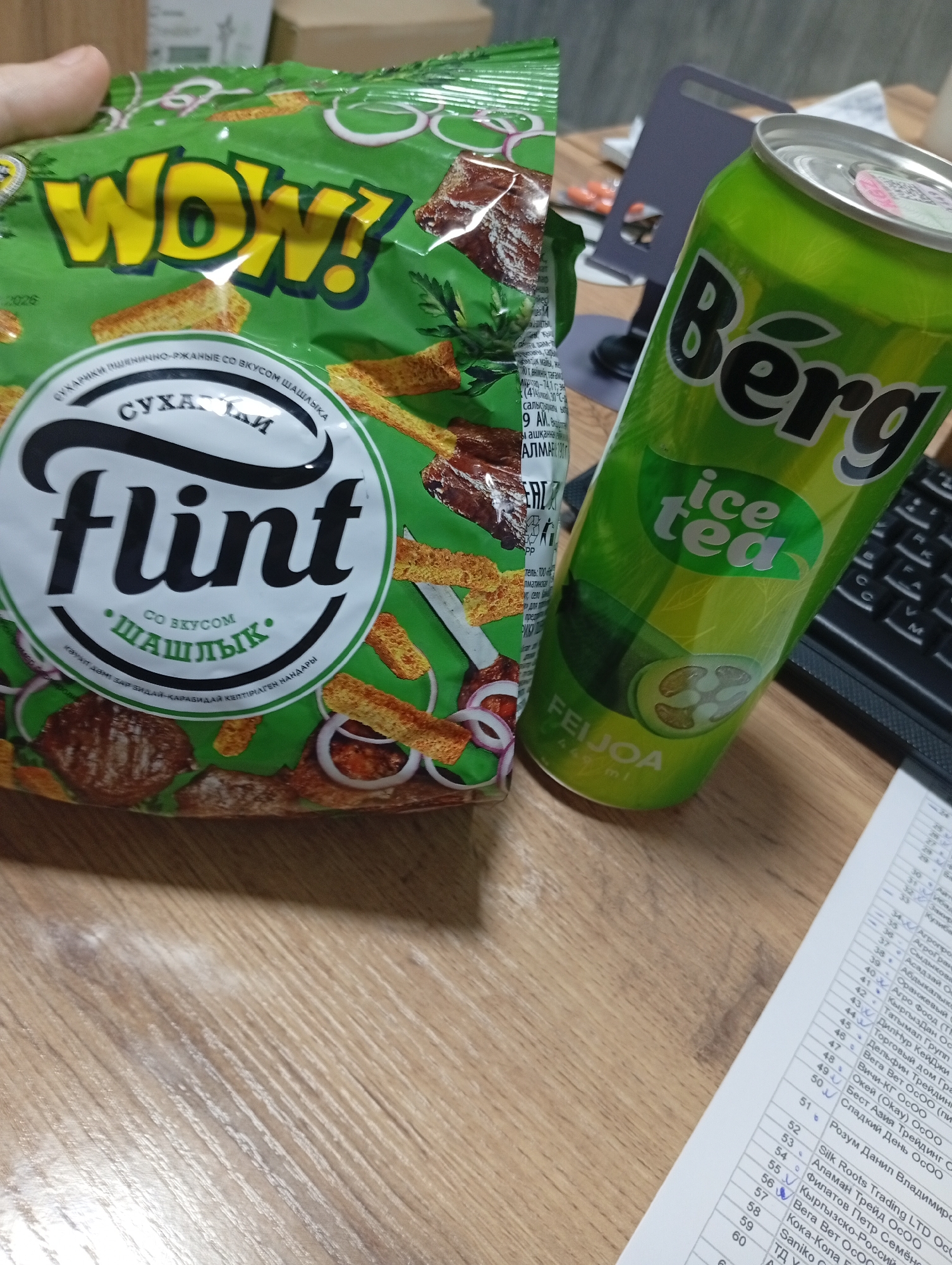 The image displays a close-up, top-down view of a wooden desk surface in what appears to be an office environment. In the foreground and slightly left of center, a green plastic bag of "Flint" brand croutons with a "Shashlyk" flavor is visible, held partially by a person's thumb and index finger. To the right of the bag, a bright green aluminum can of "Berg" brand feijoa-flavored ice tea is prominently featured, leaning against an unseen object. The background shows a keyboard, a portion of a purple tablet stand, and a sheet of paper with a list of items and numbers. The overall lighting is bright, suggesting an indoor setting, likely during daylight hours. The image does not provide any specific landmarks or visual cues to indicate its location within Osh City, Kyrgyzstan.