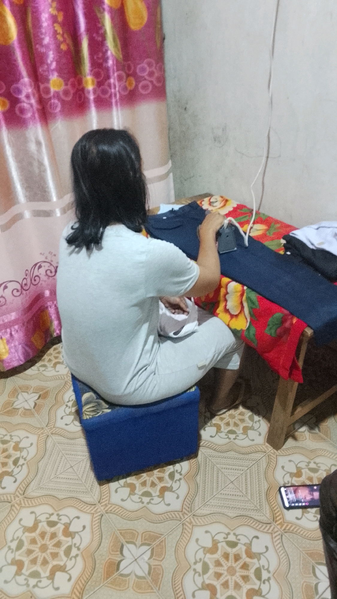 A person is seen from behind, sitting on a blue stool and ironing clothes. They are wearing a light gray t-shirt and light gray pants. The ironing is taking place on a wooden table covered with a red cloth featuring a floral pattern of red, yellow, and green flowers. A dark blue pair of pants and a white shirt are also on the table, suggesting they are being ironed. In the background, a pink and yellow patterned curtain is visible. The floor is tiled with a decorative pattern. A smartphone is lying on the floor to the right of the table. The scene appears to be indoors, in a room with light-colored walls. There is no discernible indication of the city of Mulyo Haji, Indonesia, within the image itself.