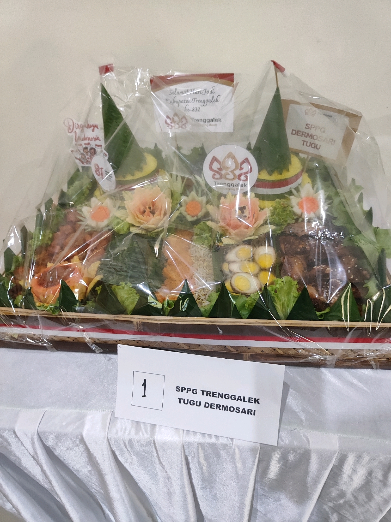 The image shows a large, elaborately decorated food arrangement, likely a tumpeng, presented on a woven platter and covered in clear plastic. The tumpeng is conical in shape and features multiple tiers of food, including rice, meats, vegetables, and hard-boiled eggs. Decorative elements, such as carved fruits resembling flowers, are interspersed throughout. Several small flags and labels are attached to the arrangement.
One prominent label, positioned in the lower right corner of the image on a white card, displays the number "1" in a square box, followed by the text "SPPG TRENGGALEK TUGU DERMOSARI". This suggests the food arrangement is associated with a specific location or organization within Trenggalek, Indonesia.
Another label, near the top center of the tumpeng, reads "Selamat Hari Jadi Ke-832 Kabupaten Trenggalek" (Happy 832nd Anniversary of Trenggalek Regency). This indicates the context of the presentation is a celebration of Trenggalek's anniversary.
A third label, on the left side of the tumpeng, says "Dirgahayu Indonesia" (Long Live Indonesia), suggesting a patriotic theme. A circular logo with "Trenggalek" written within it is also visible.
The tumpeng is placed on a white tablecloth with a pleated texture, which drapes over the edge of a table. The background is a plain, light-colored wall. The overall scene appears to be an indoor event or display, possibly in a community hall or an office setting. The lighting is even, suggesting a well-lit indoor environment. No people are directly visible in the image, but the presence of the tumpeng and the celebratory labels implies a gathering or event is taking place.