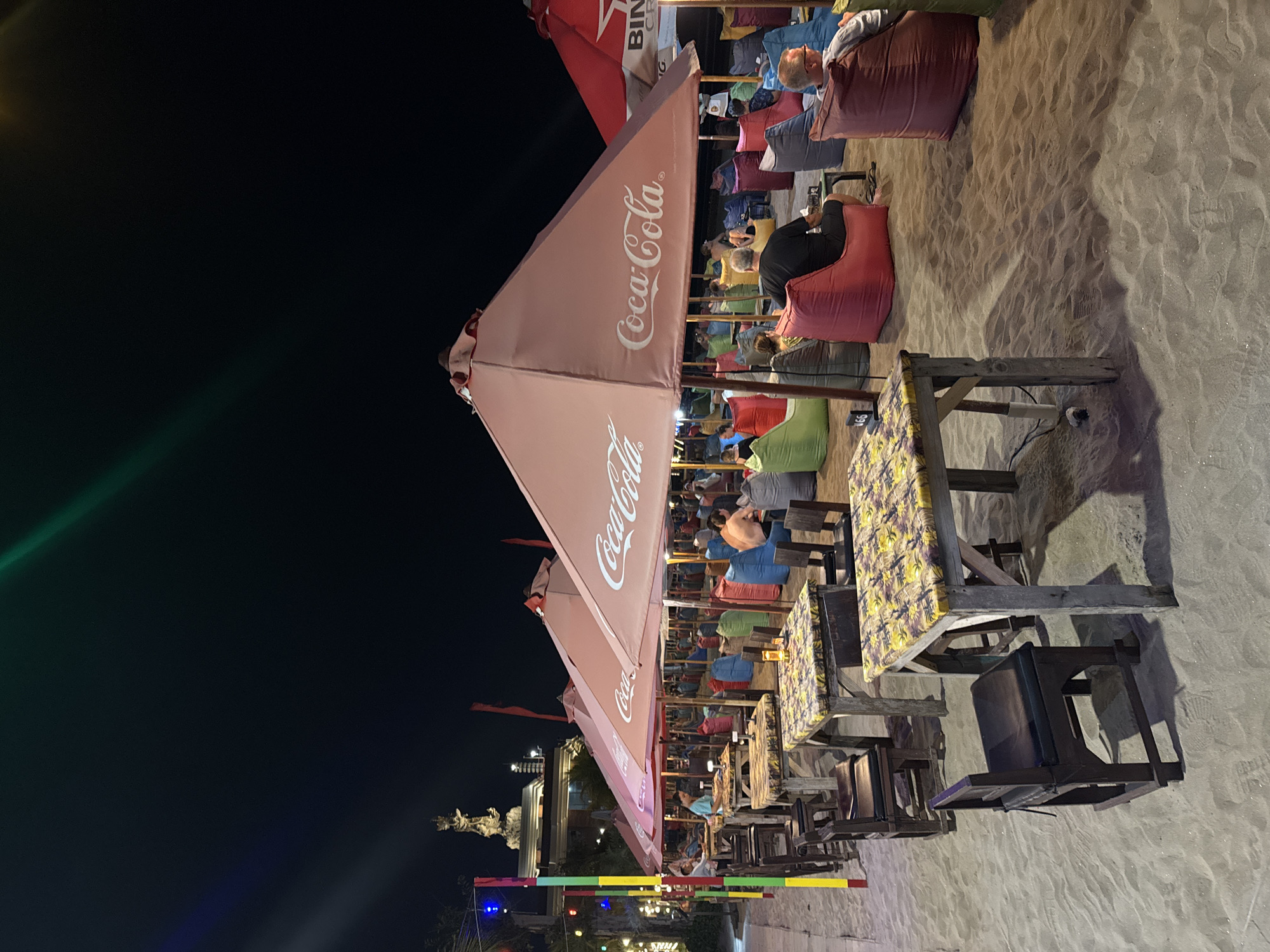 An outdoor beachside restaurant in Kuta, Indonesia is captured at night. The scene is illuminated by overhead lights and string lights, creating a warm ambiance. Numerous tables with checkered tablecloths and wooden chairs are set up on the sandy beach. Large, pink umbrellas, some displaying the Coca-Cola logo, provide shade. Beanbag chairs in various colors are arranged around some of the seating areas, occupied by patrons. In the background, a statue of a deity is visible atop a building, along with other structures and the dark night sky. The overall atmosphere suggests a relaxed and lively dining experience on the beach.