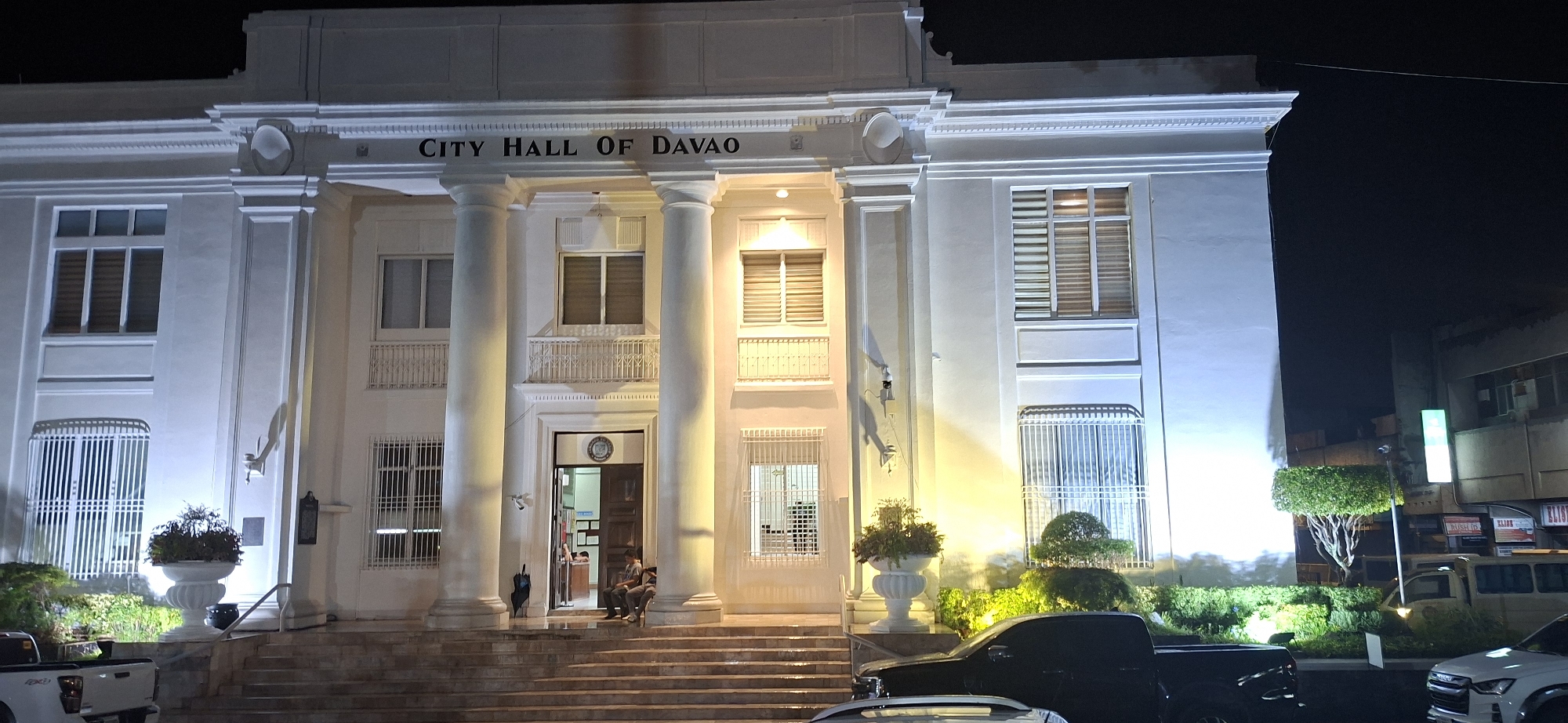 The image shows the facade of the City Hall of Davao, a white, neoclassical building at night. The building features a prominent portico with large columns and a pediment, above which the words "CITY HALL OF DAVAO" are inscribed. There are several windows with louvered blinds and decorative railings on the upper floors. A wide set of stairs leads up to the main entrance, where two individuals are seated. A large potted plant is visible on the landing. The building is illuminated by artificial lights, casting shadows and highlighting its architectural features. In the foreground, parts of vehicles, including a black pickup truck, are visible. The surrounding environment is dark, with the night sky above and some greenery and other buildings in the background.
