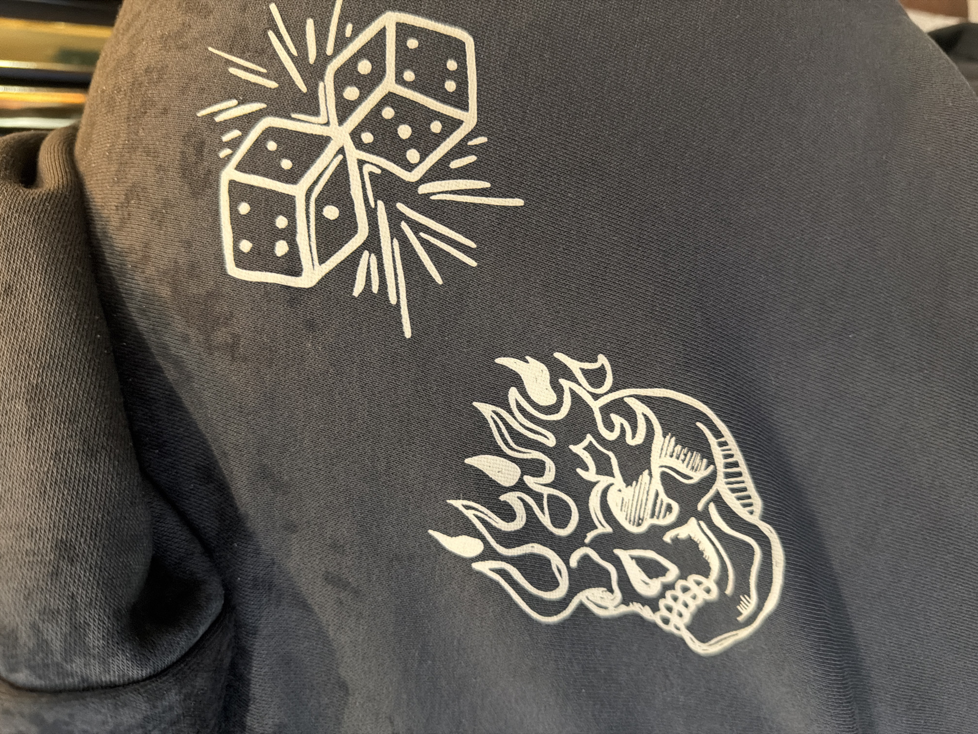 The image shows a close-up of a dark gray hooded sweatshirt. Printed in white on the upper left is an image of two dice with radiating lines, suggesting movement or impact. Below the dice, on the lower right, is a stylized skull surrounded by flames. The background is the fabric of the sweatshirt. The location in the city of Mole Valley, United Kingdom, is not discernable from the image.