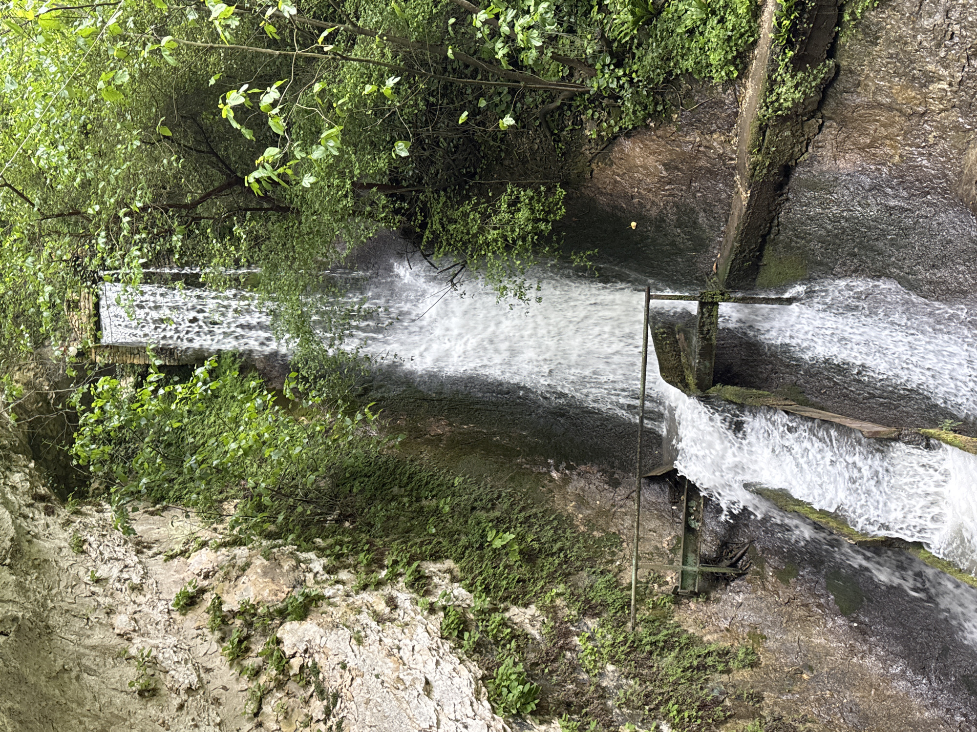 The image depicts a waterfall in Tsabal, Abkhazia. Water cascades over a man-made structure consisting of a series of stepped dams and concrete channels. The waterfall flows through a lush, green environment with trees and vegetation along the banks. The scene appears to be set within a ravine or gorge. A metal framework sits within the cascading water, and some logs also float on the water. The rock face is partially covered in green moss and vegetation. The water is turbulent and whitewater.